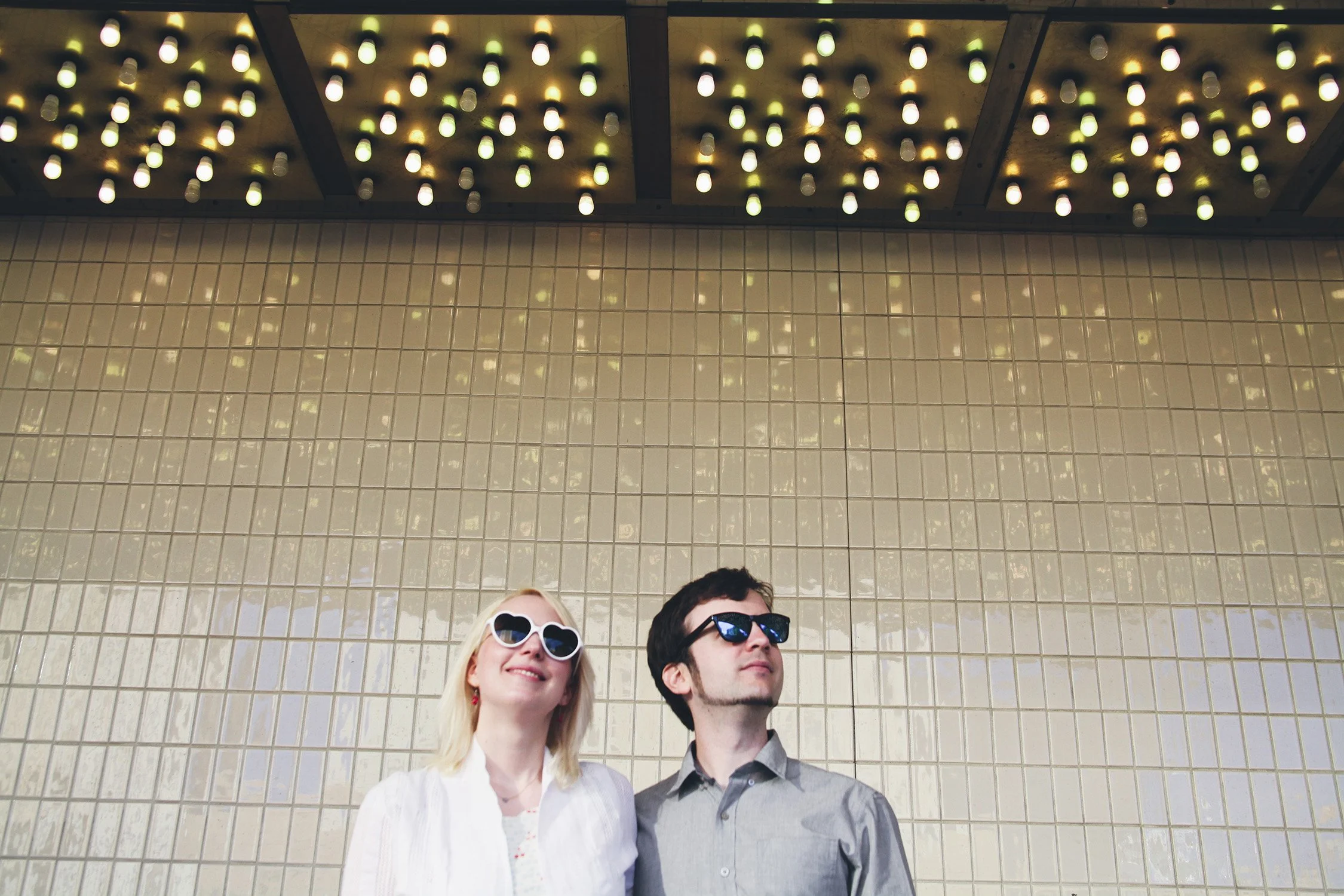 Engaged couple standing in front of white subway tile wall with Vegas glitter lights above, both wearing sunglasses and smiling while looking off camera