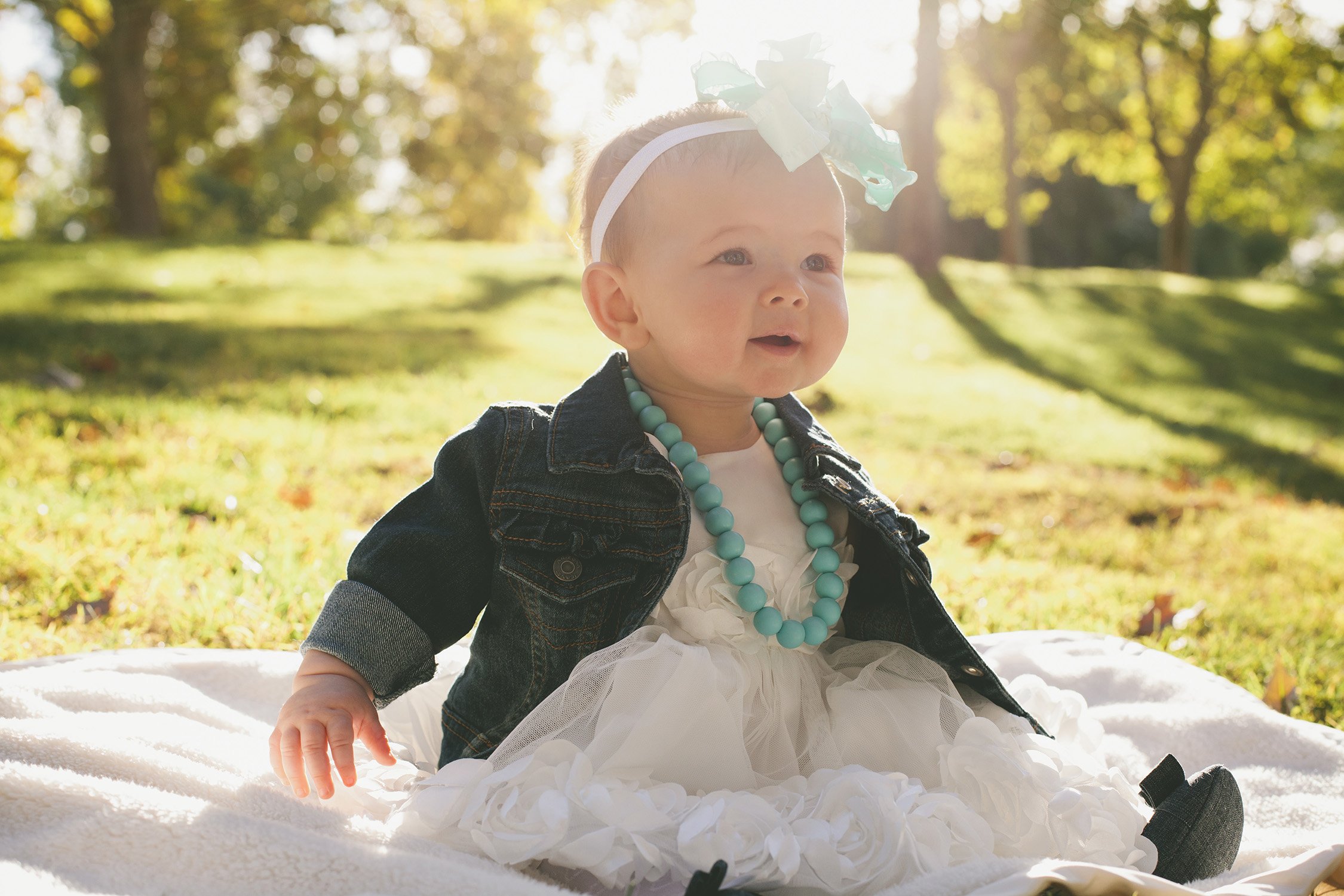 A young baby is sitting up on a blanket on the grass with trees and sunlight in the background. She is wearing a big cute light blue and white bow in her on head smiling off camera