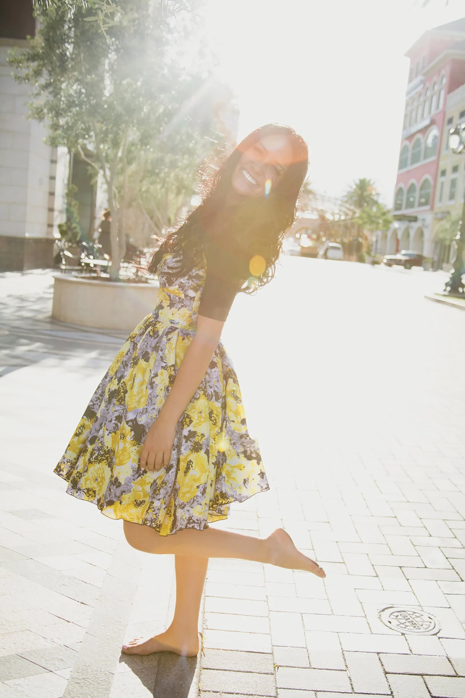 Standing on a stone road with the bright sun behind her, high school senior, Ashley is wearing a bright yellow flower dress and is standing with one leg up and leaning back smiling at the camera.