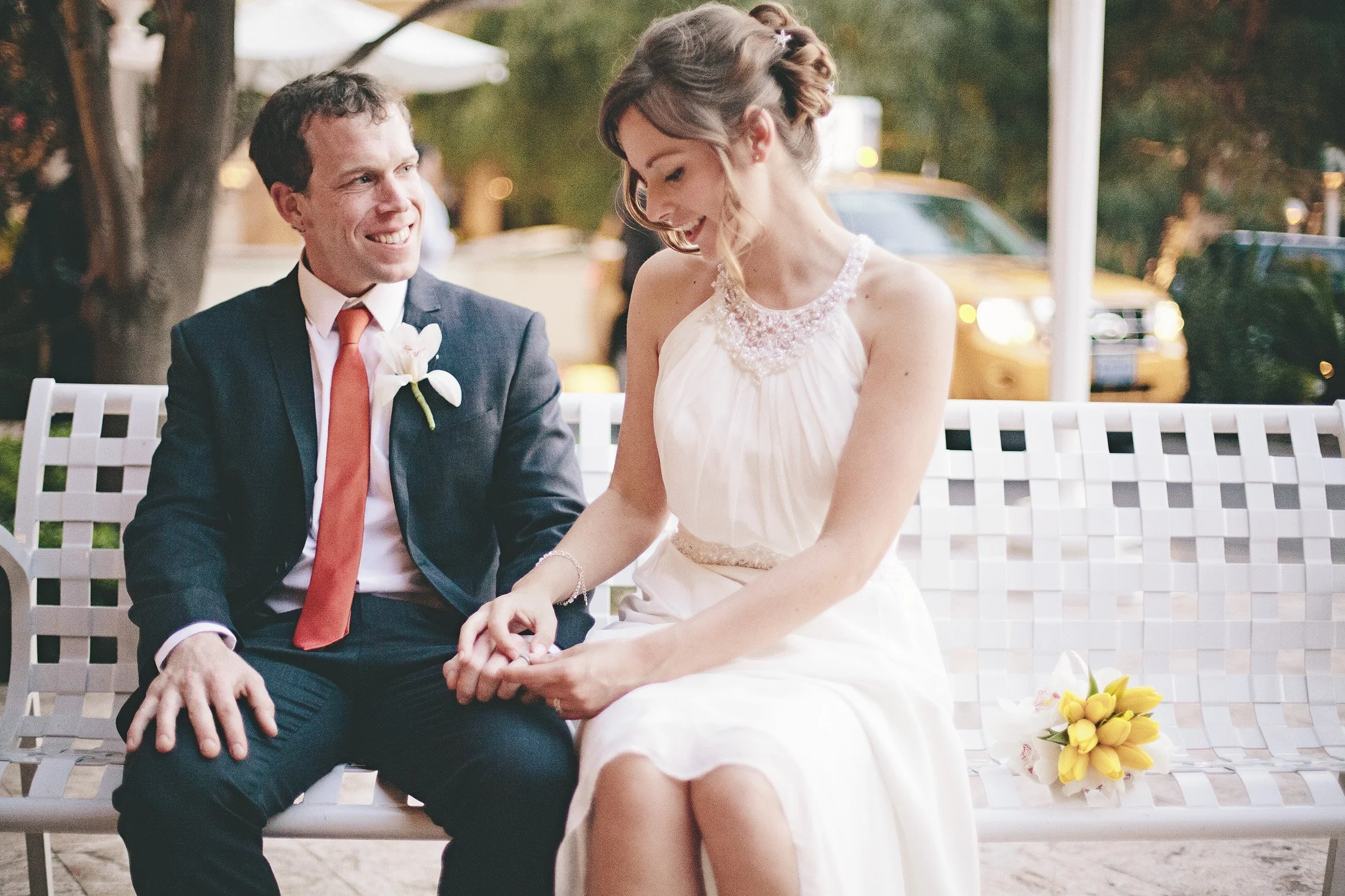 Newly weds sitting on a bench, groom is smiling at the bride, the bride is looking at the ring on his finger, the bouquet of flowers sitting on the bench and a taxi driving by in the background