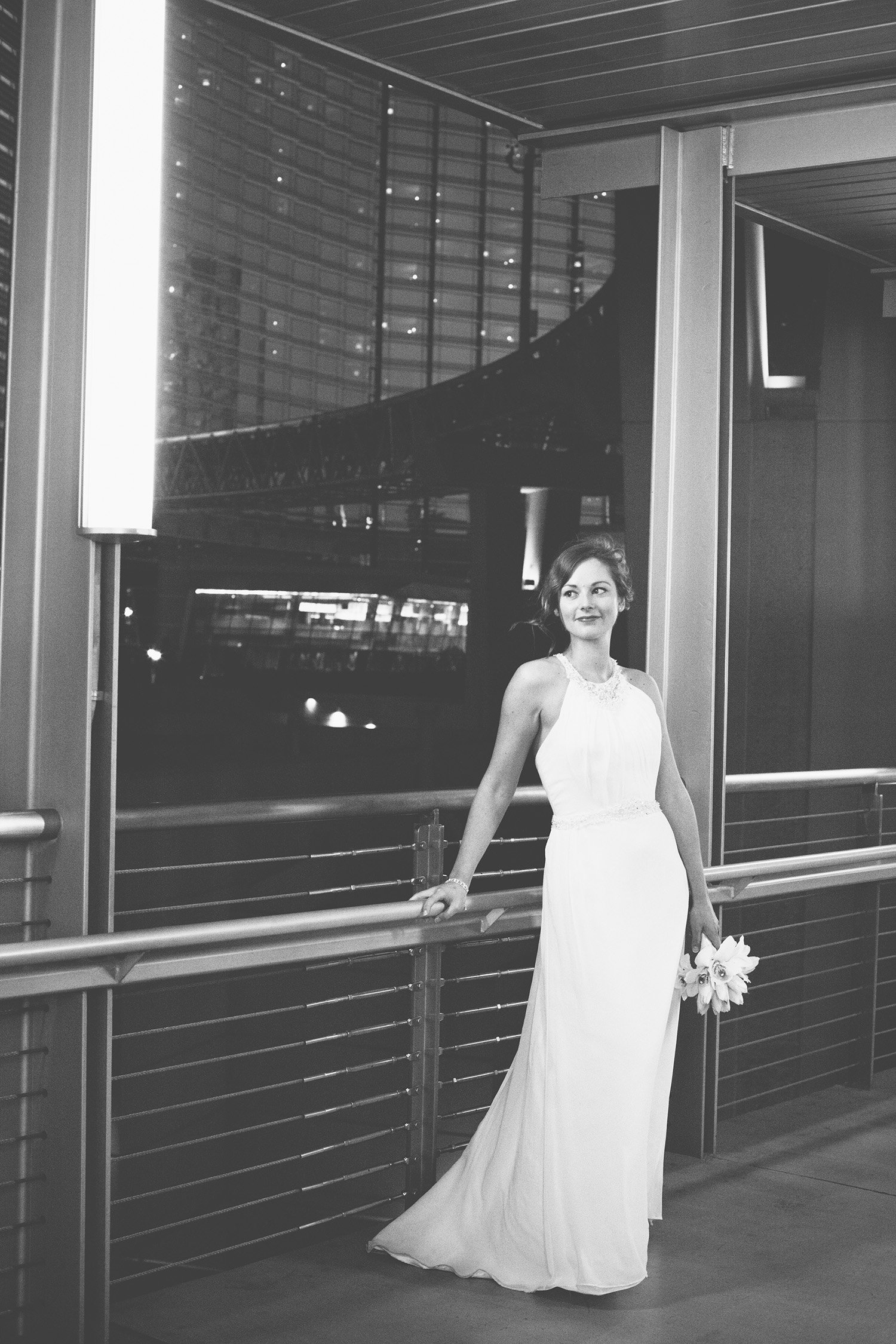 a black and white image of a bride holding her bouquet in one hand, leanding and holding a railing in the other hand with a tall glass hotel in the background