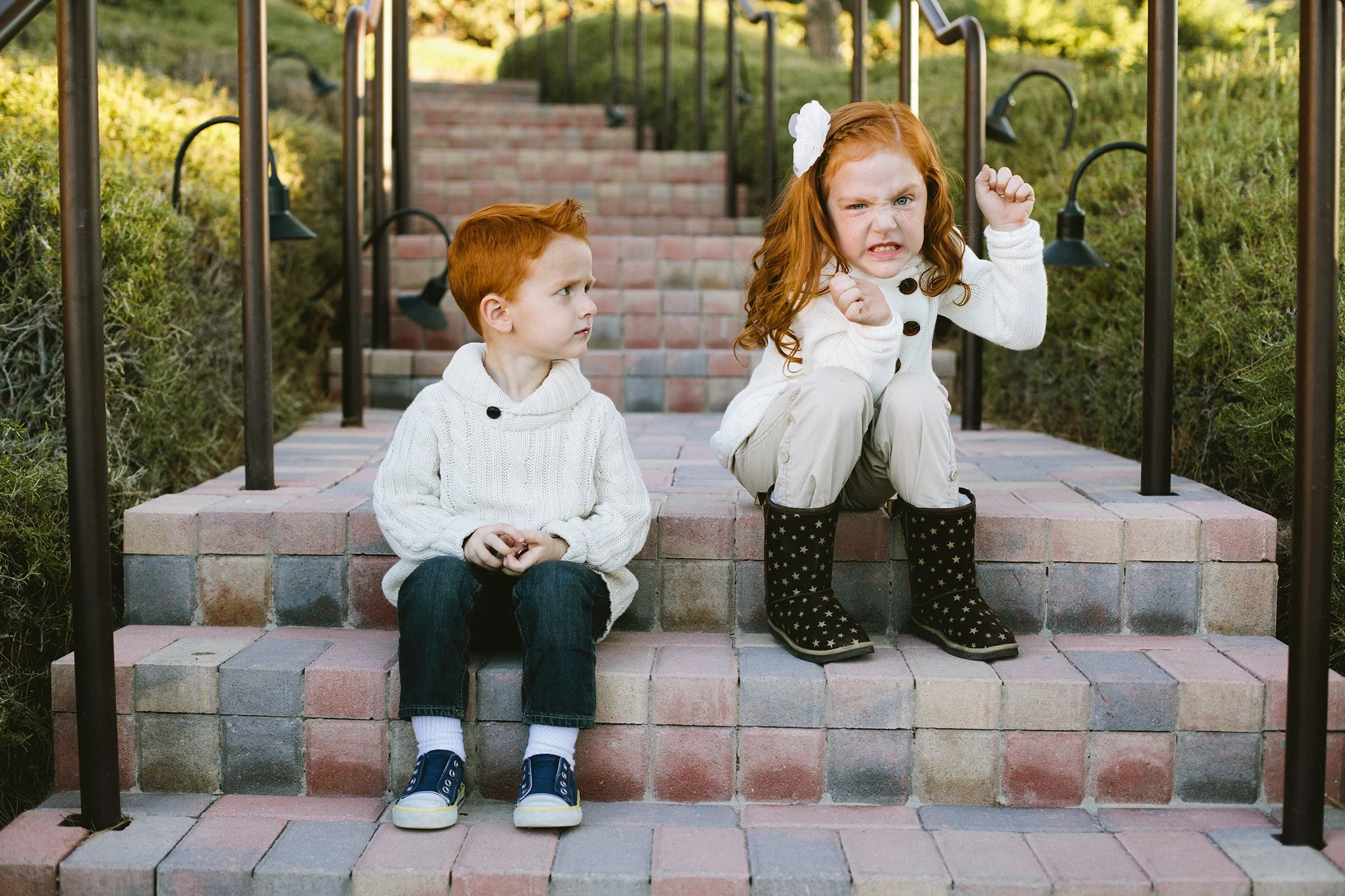 on a brick stair path, two redheaded young siblings on sitting on steps in the foreground. The older sister is making a funny angry face at the camera and the younger brother is looking confused at the sister.