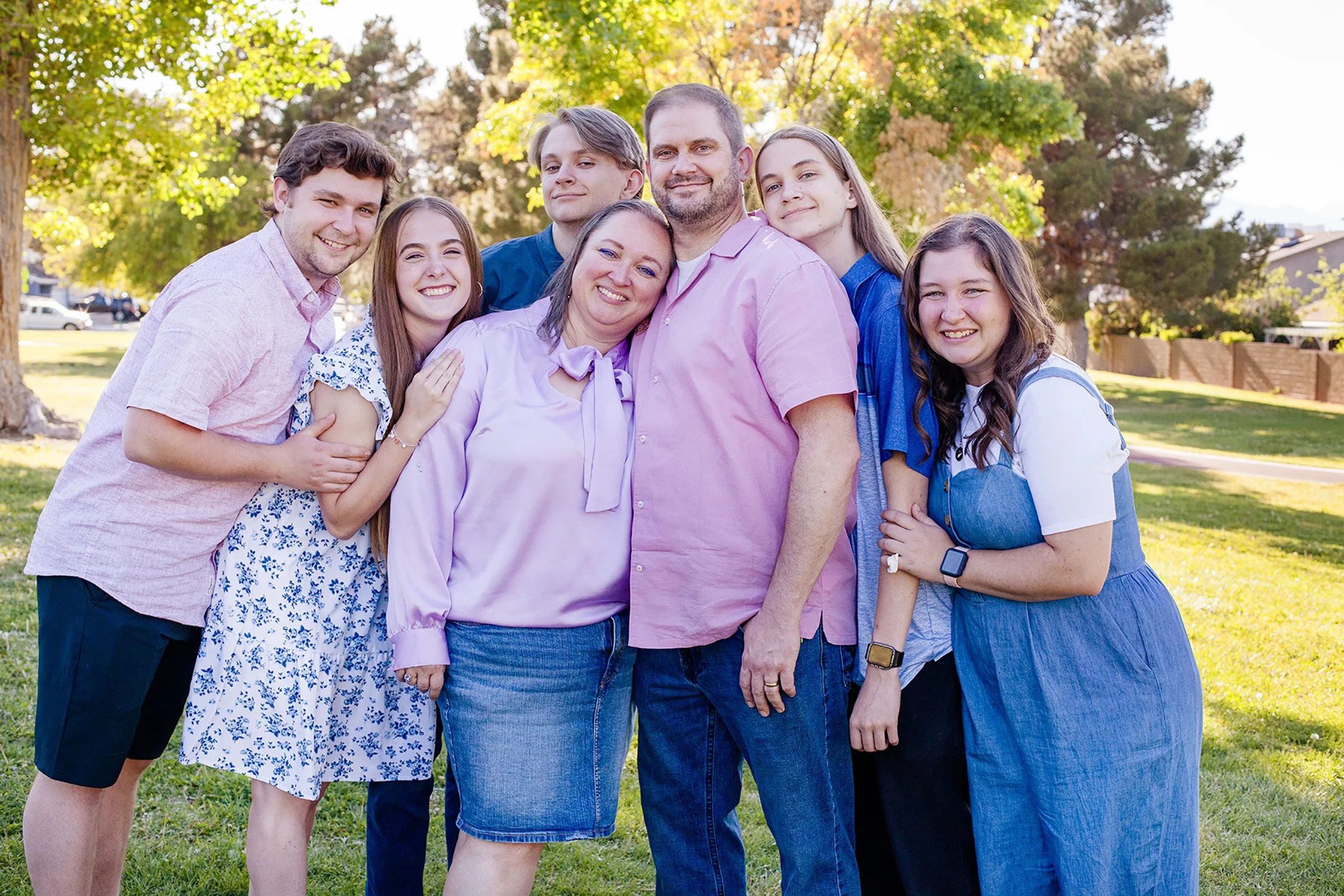 a family of seven, hugging together around mom and dad. All the kids are adults or older teenager age, smiling at the camera, with the park's trees and grass in the background on a sunny day.
