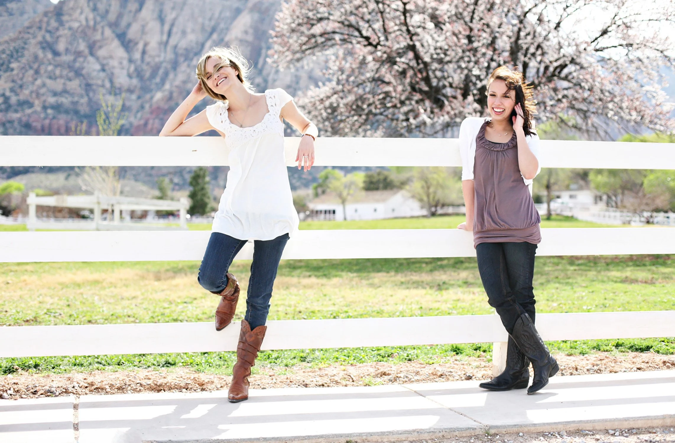 standing against a white wood fence, two high school seniors are looking away from the camera smiling, there is a big beautiful tree with white and pink blossoms behind them including a green pasture and mountains.