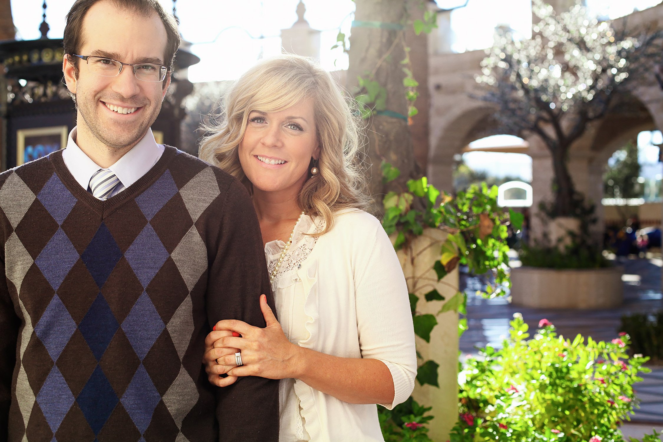 A couple standing close together for engagement photos, the sun is shining behind them, she is holding on to his arm and they are both smiling at the camera