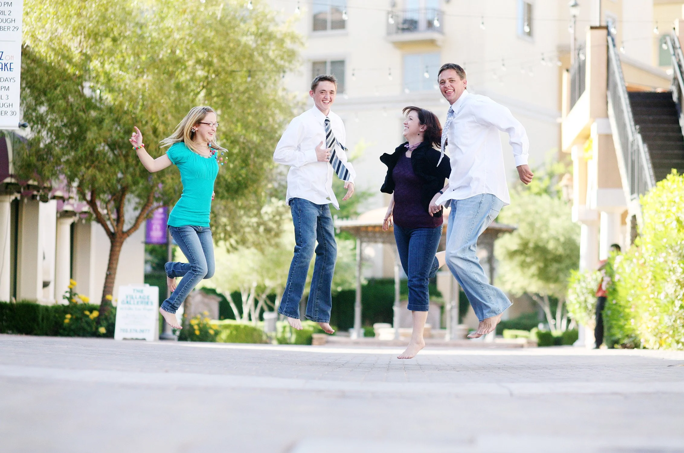 A family of four, two young teenagers and a mom and dad are in the middle of a wide quiet brick pathway shopping area jumping in the air together, smiling at the camera or at each other