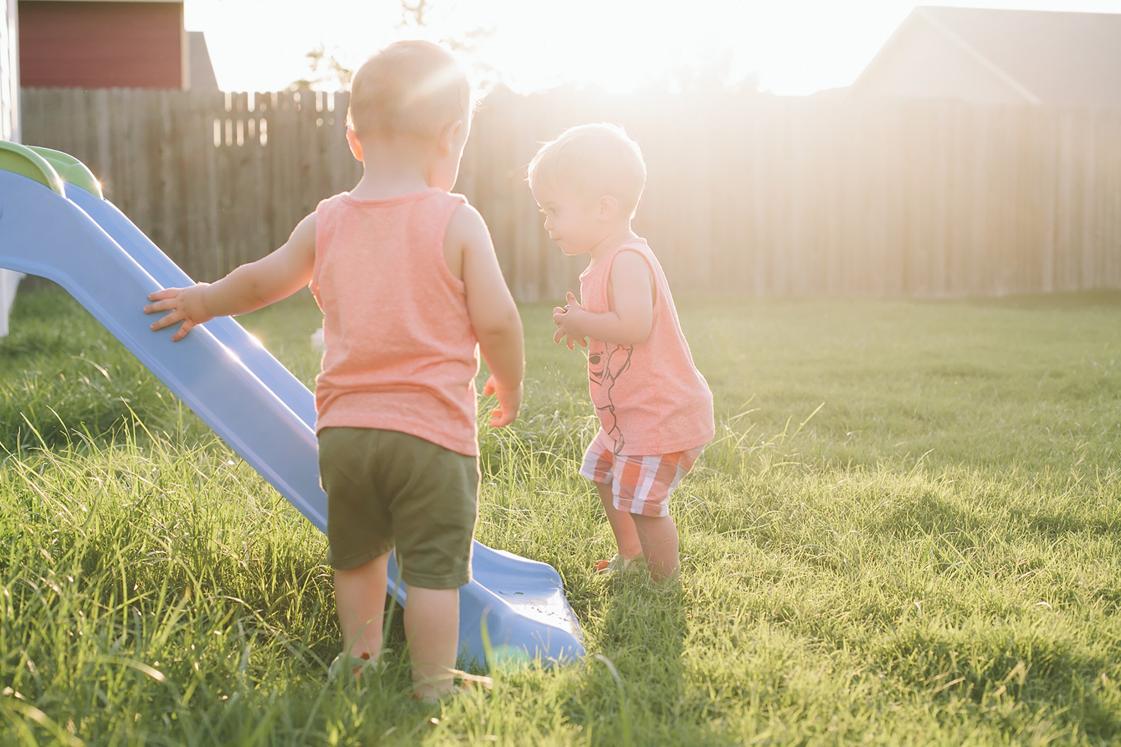 two young brothers around 2 years old are standing in the grass around the bottom of a small kid slide, the sun is behind them and they are in a backyard with a tall wood fence in the background