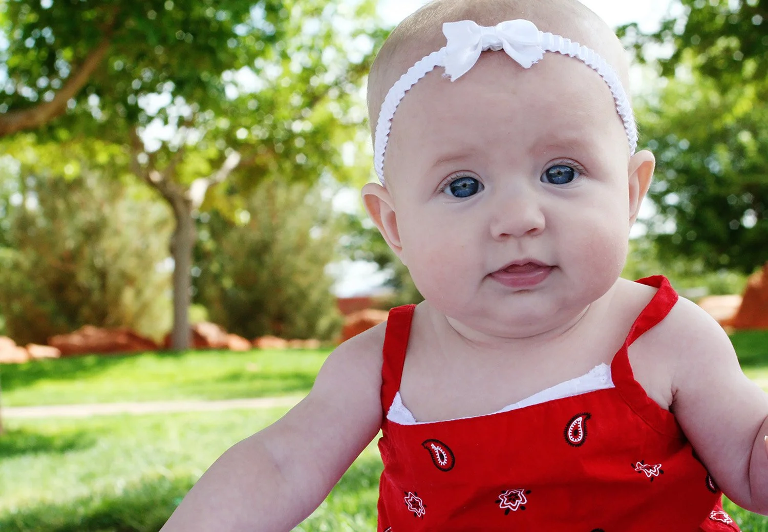 a baby at a park, looking directly into the camera, wearing a red tank top and white bow in her hair