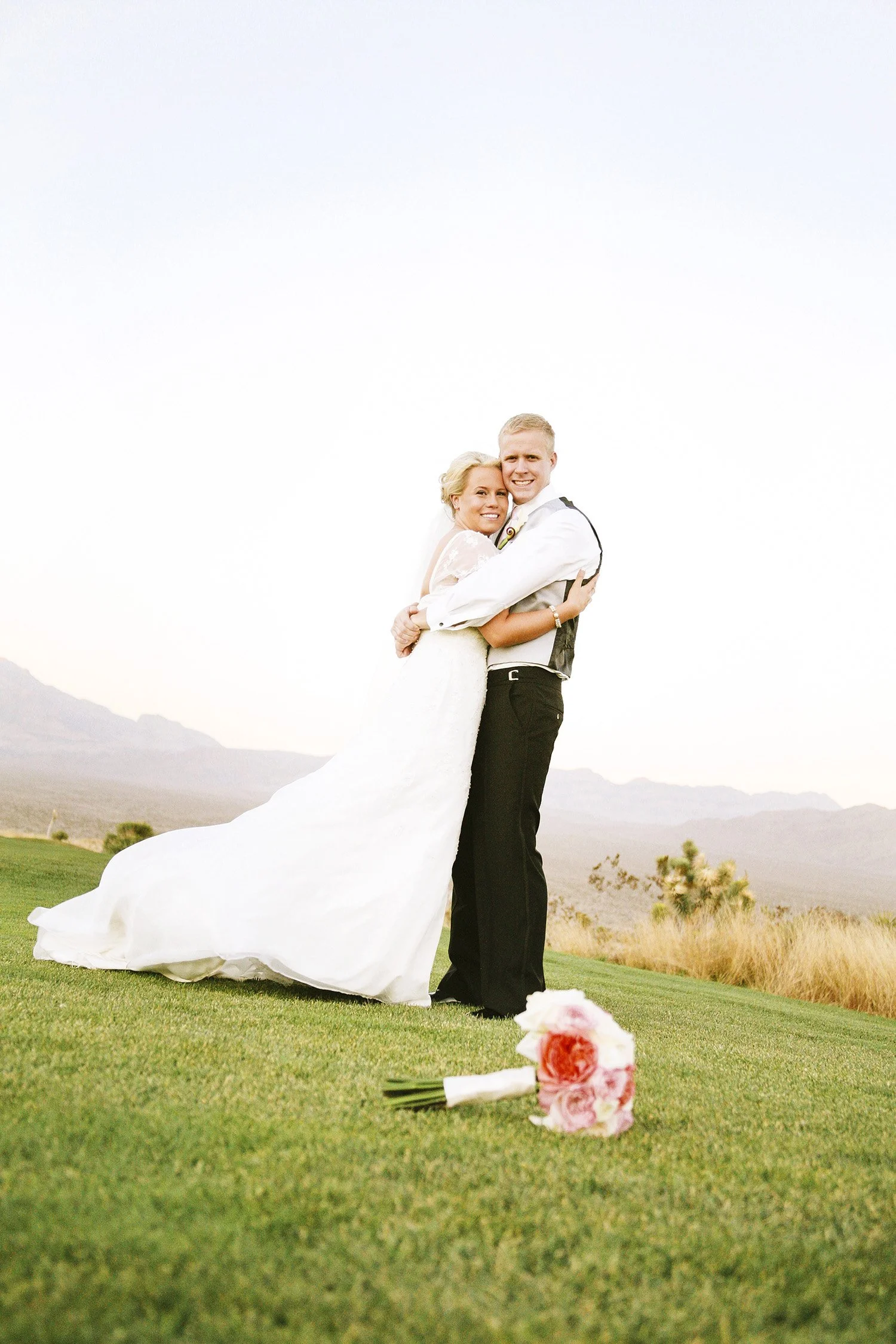 a newly wed couple hugging, smiling and looking at the camera, standing on grass with a bouquet in the foreground and a purple mountain range in the background