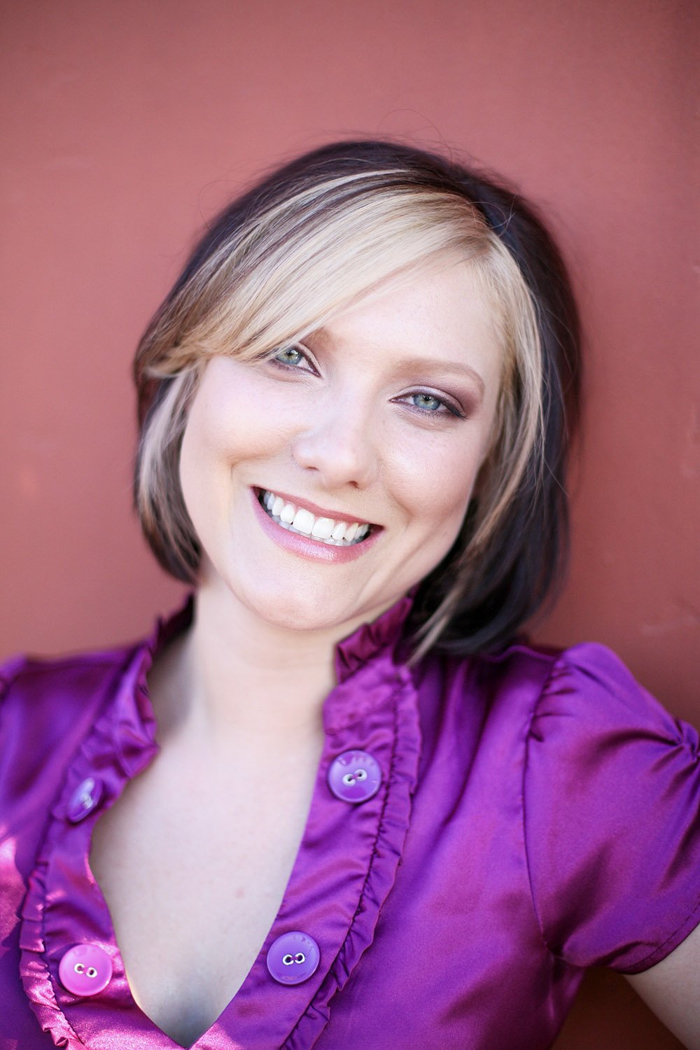 An up close image of a young woman smiling at the camera, sitting against a pink wall, wearing a purple shirt, with bright blue eyes
