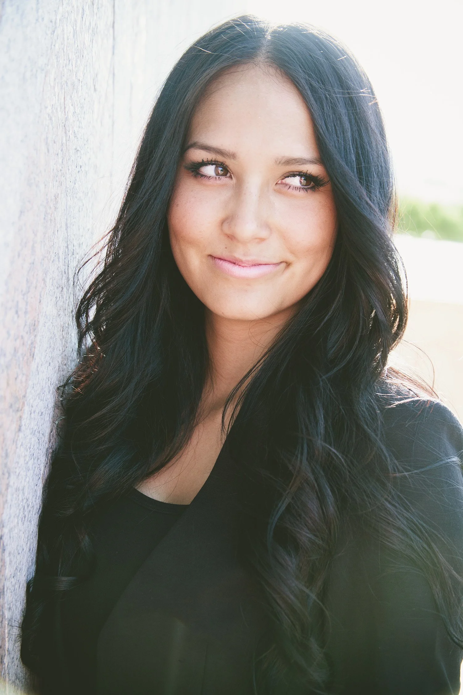 One shoulder leaning against a wall, high school senior, Ashley, is wearing all black and looking off camera as she gives a cute smirk. This is a close up image.