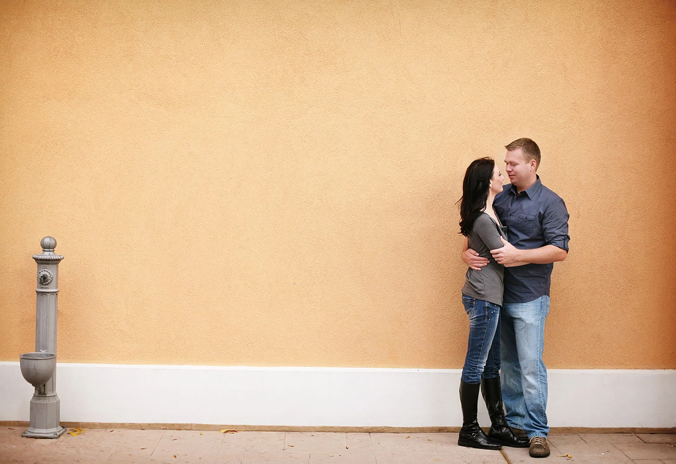 A minimal photos of an engaged couple, standing by a taupe wall, holding each other close looking at each other, with an Italian water spout on the other side of the photo