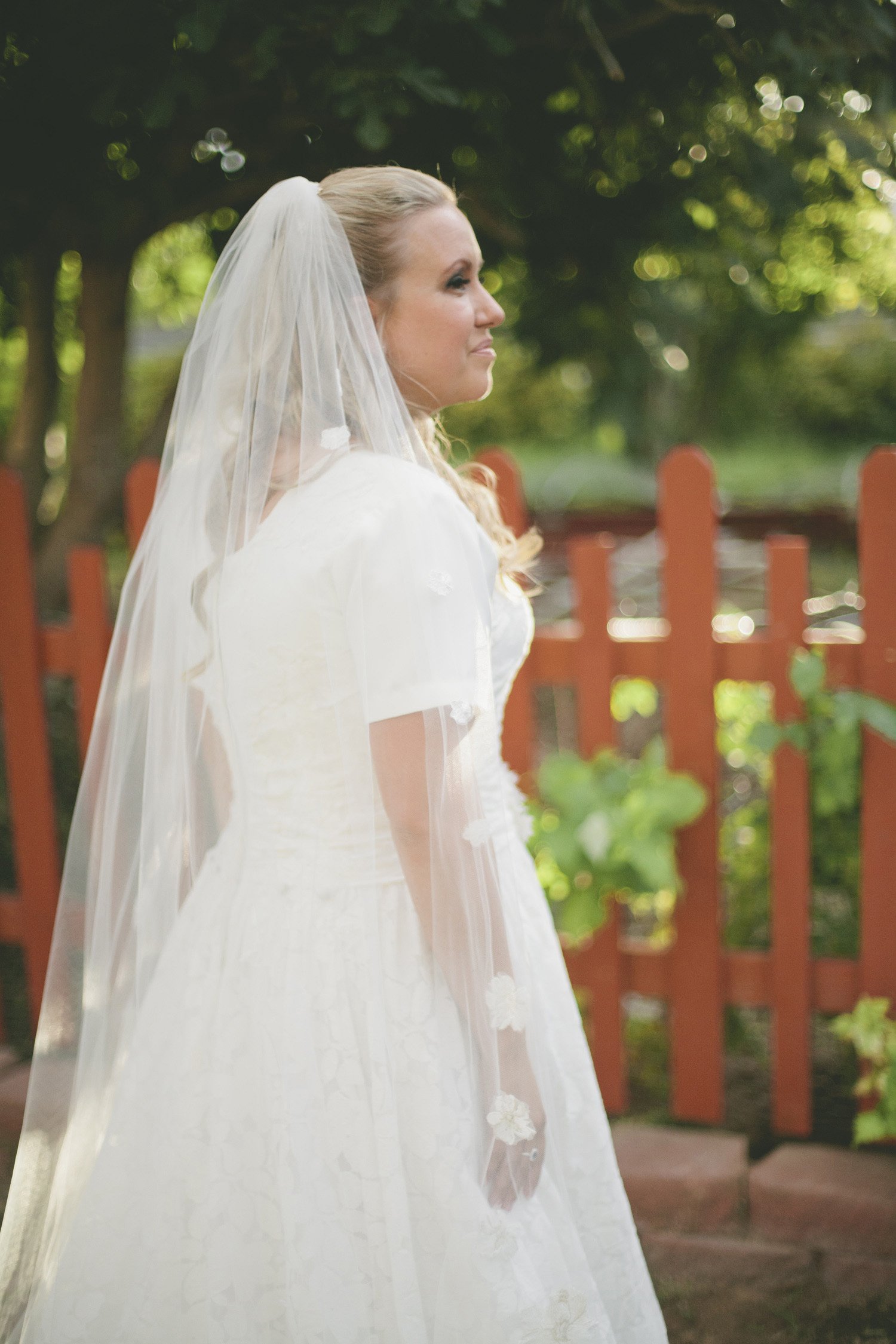 A bride in her gown, face in profile, smiling away from camera, with a red picket fence, trees and garden in the background with the sun peeking through
