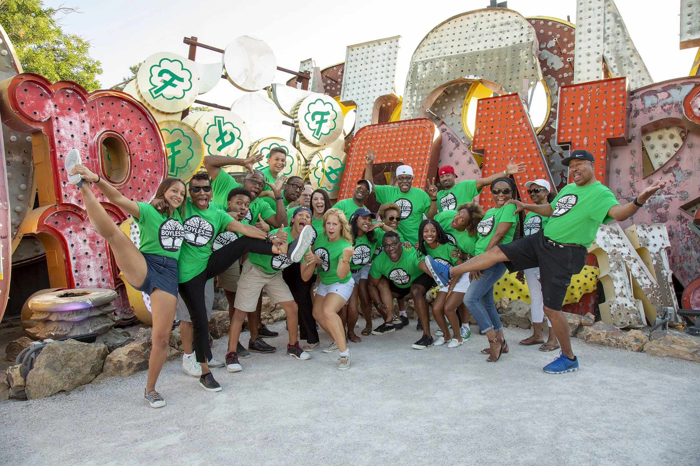A large family group are doing wacky, fun, silly posing close together, all wearing the same shirts for a family reunion. Behind them are large decommissioned neon signs. The age ranges from retired age to teenagers.