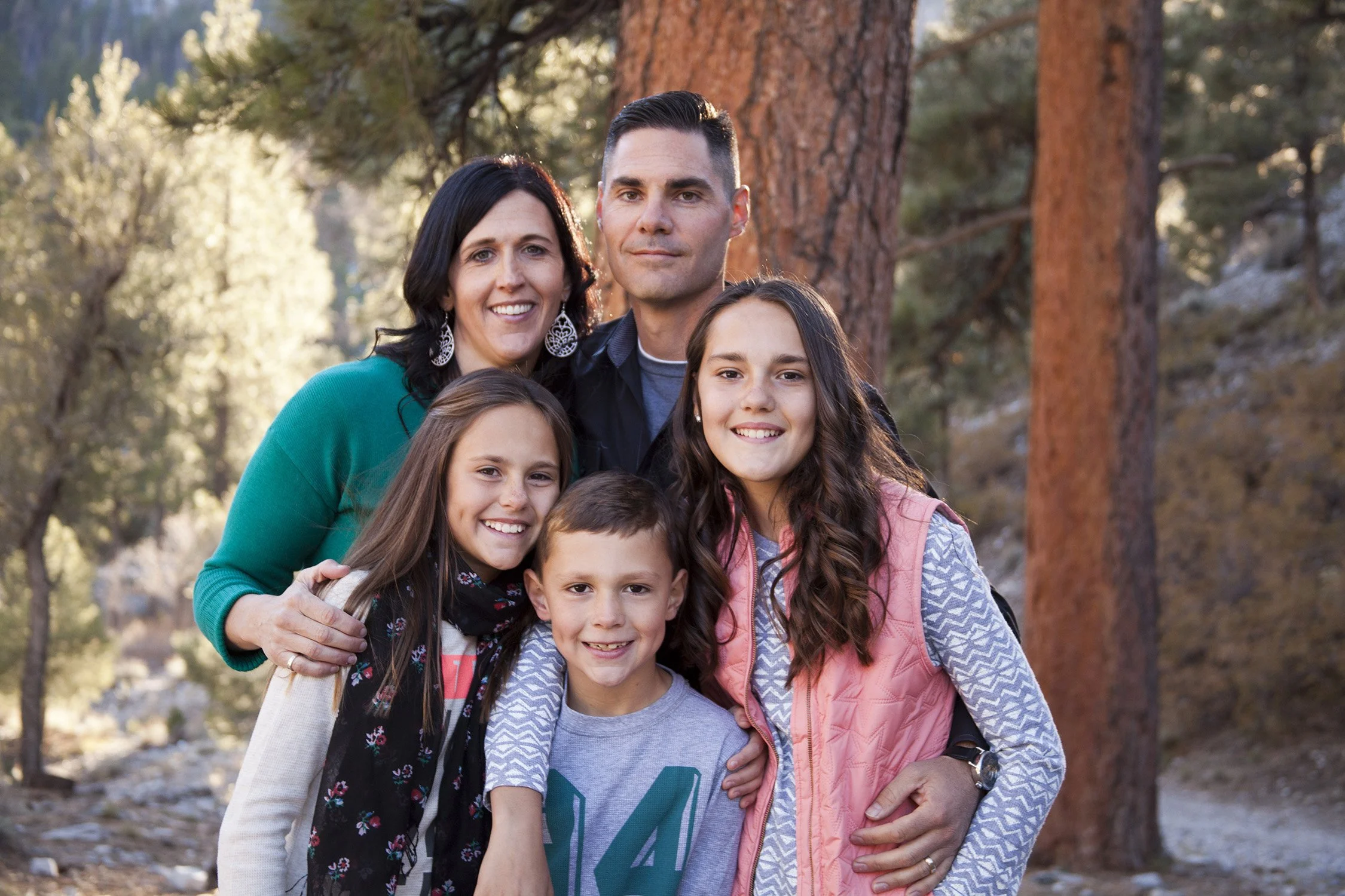A family of five are close together, in the mountain woods behind them. The mom and dad are in the back of the group, the sisters are close to the parents and younger brother in front of the group