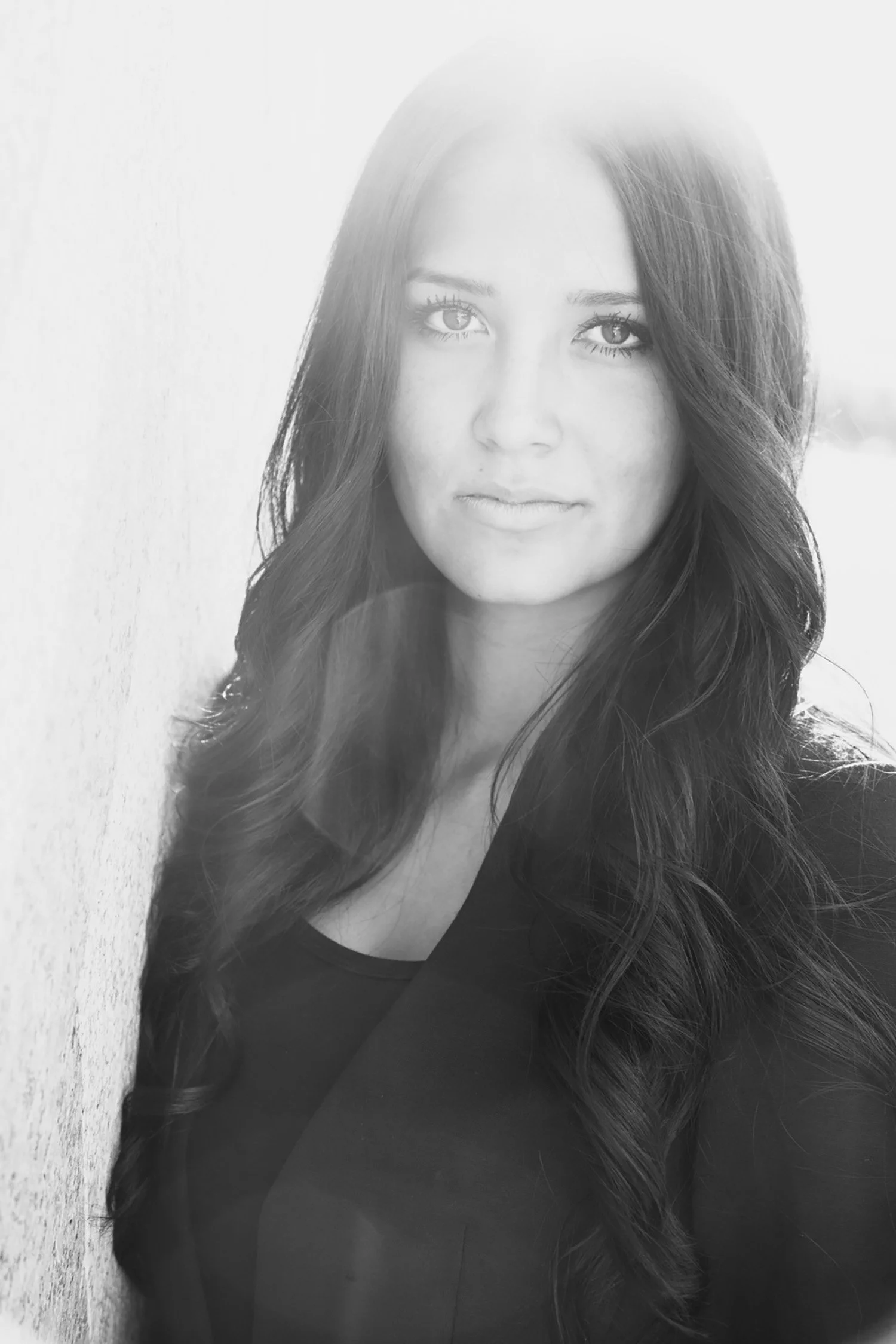A black and white image, high school senior Ashley is making a fairly serious face looking directly into the camera. There is a sun flair behind her, she is leaning on a wall.