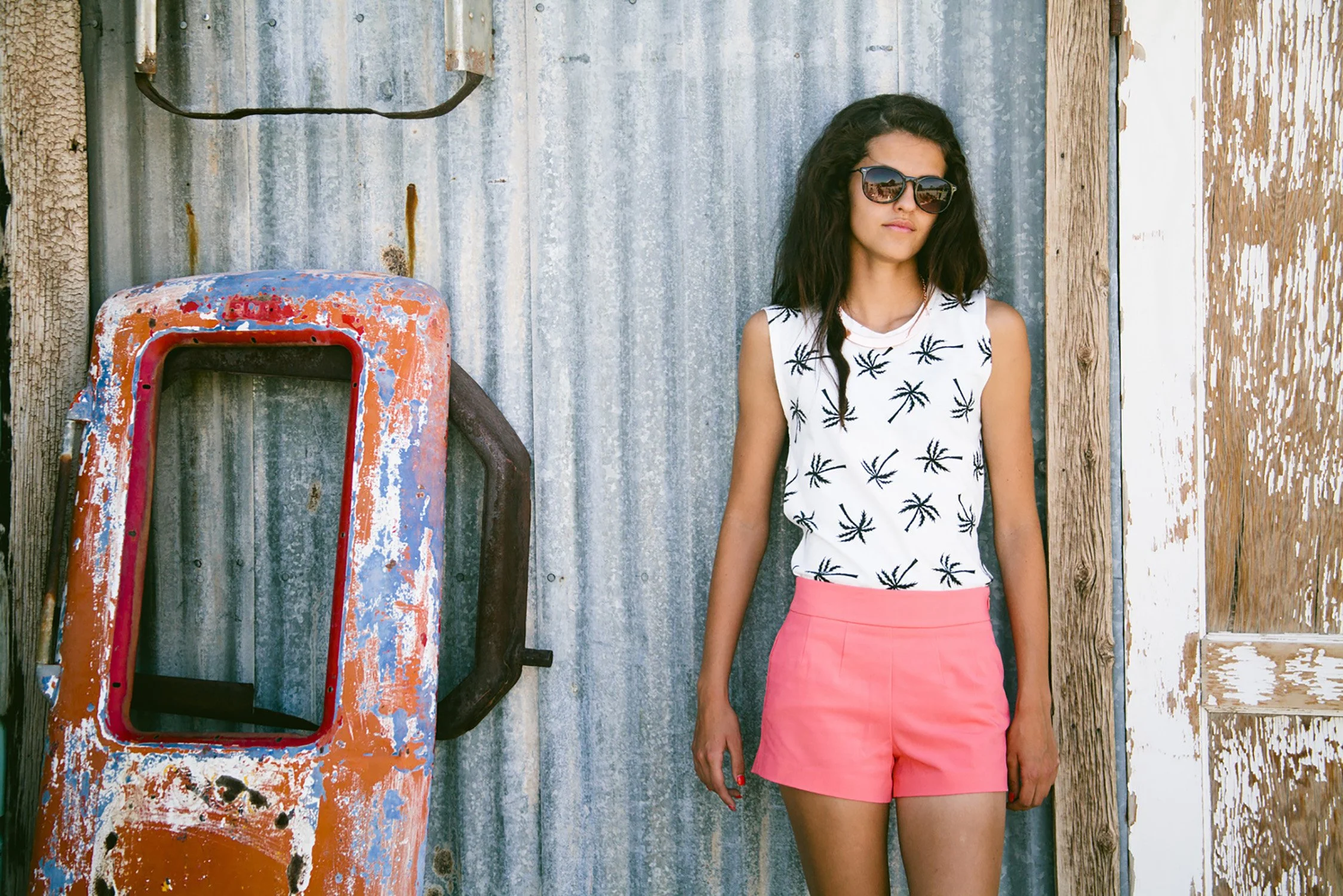 a high school senior is leaning up against a metal wall, in an old rustic area. She is wearing pink shorts and a white sleeveless shirt with palm trees and sunglasses. She is slightly smiling at the camera.