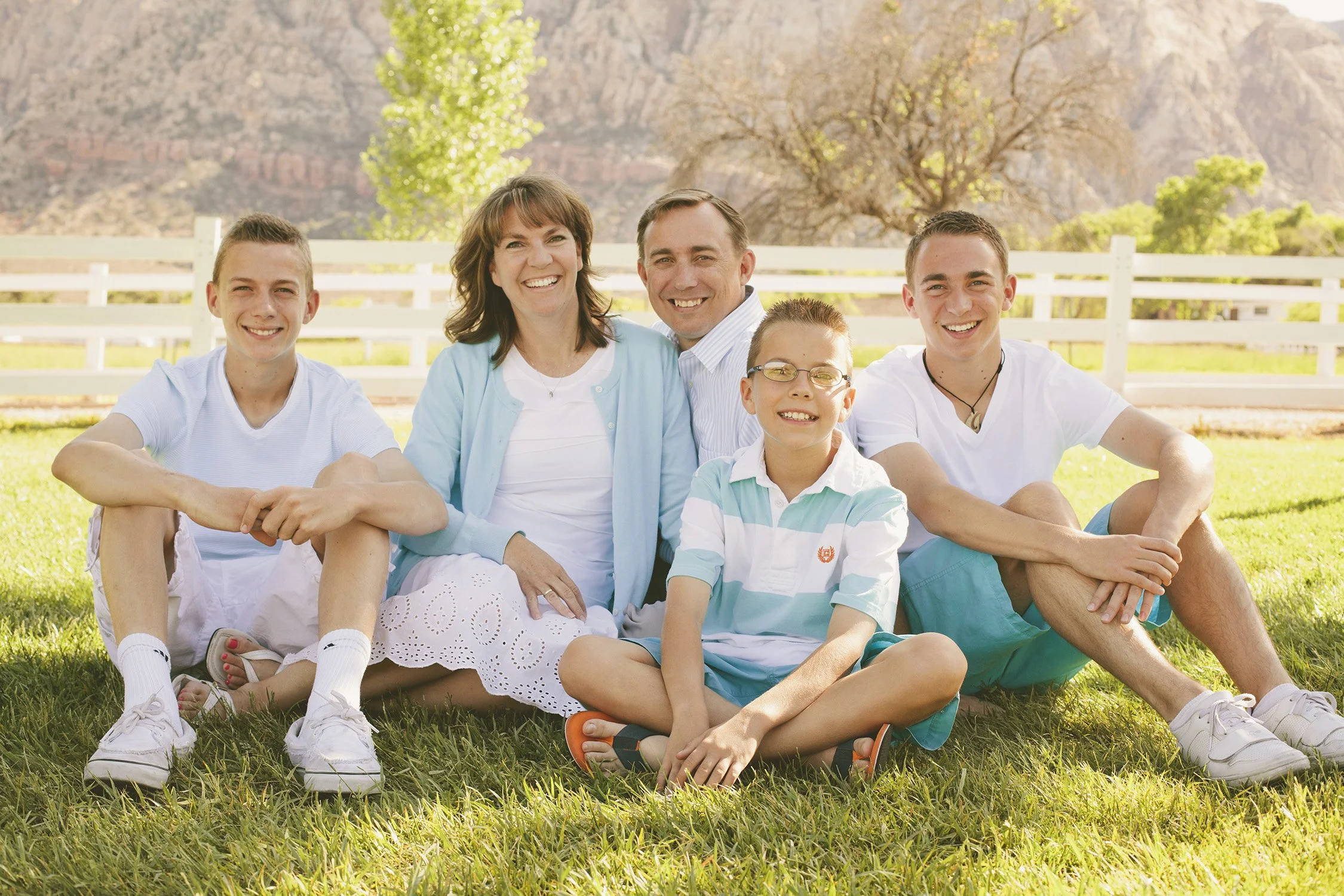 A family of five is sitting close together on grass in the shadow of a tree. The parents are the background and three sons around them, age ranging from 10 to 19 years old. They are smiling at the camera, the background has a mountain in the distance