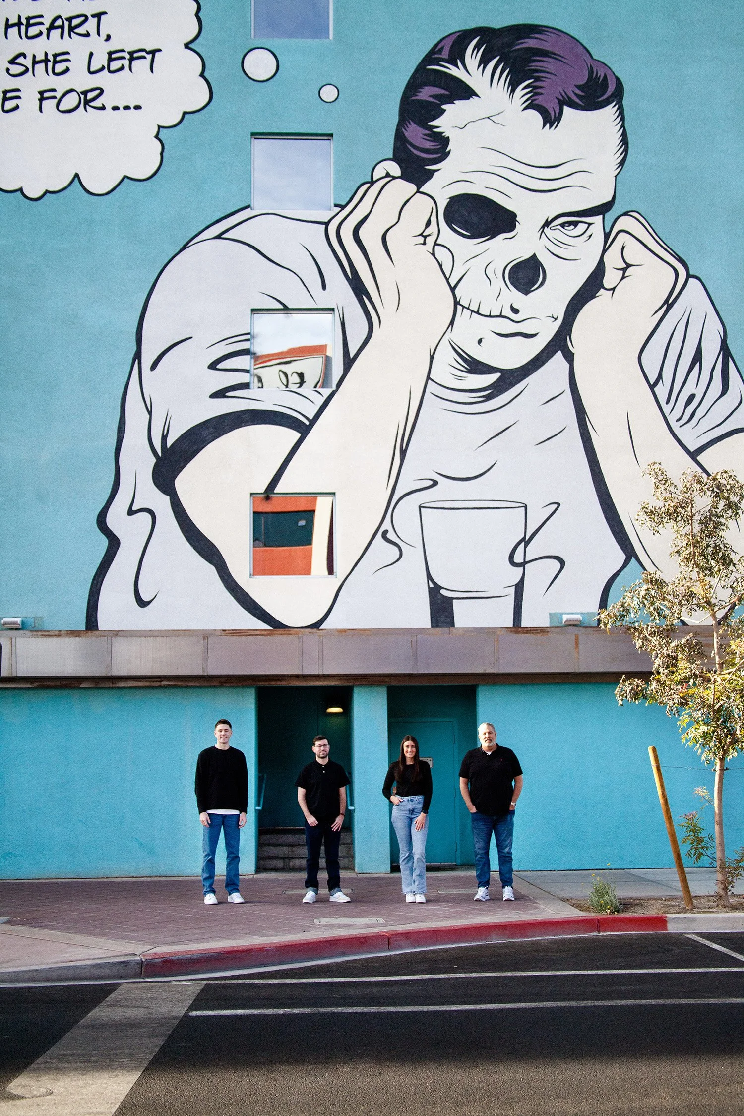 A family of four are standing together but about two feed from each other in front of a large building with a mural of a guy sulking above them