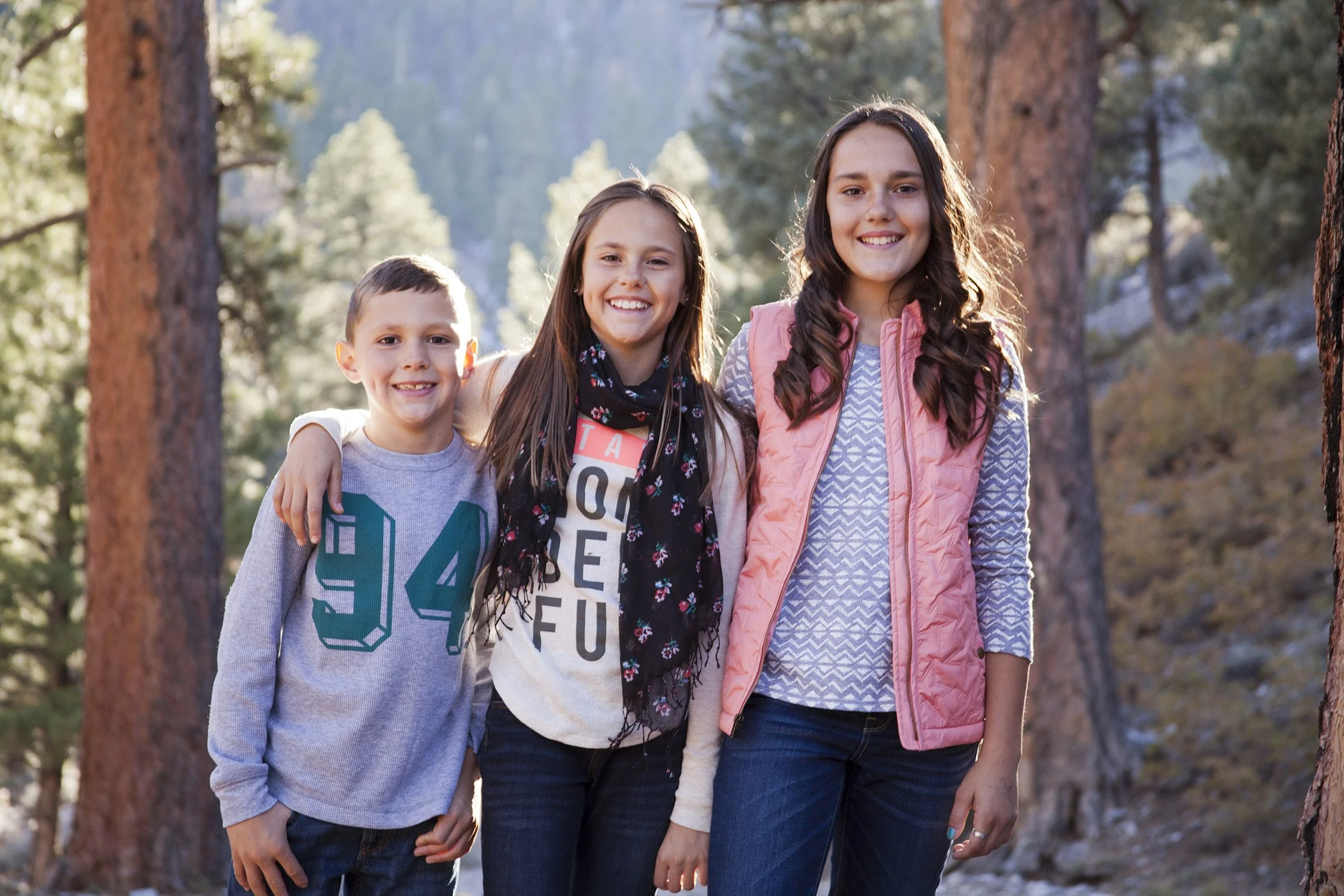 three siblings are standing in height ascension with their arms around each other. The background shows they are in the mountains among pine tress with the sunlight behind them 