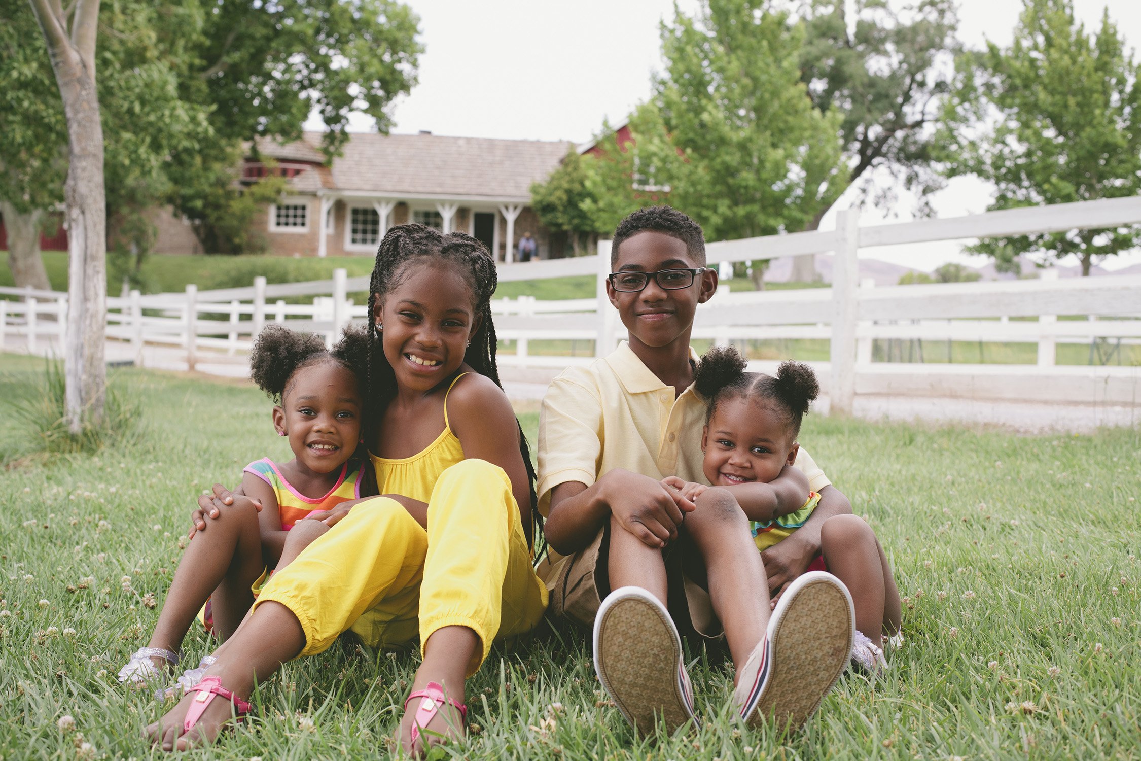 Four siblings are sitting in the grass, the two older siblings, boy and girl in the middle and the two younger girl siblings on either side of them, all are smiling at the camera. The background shows a white fence, trees and ranch style home on a sm