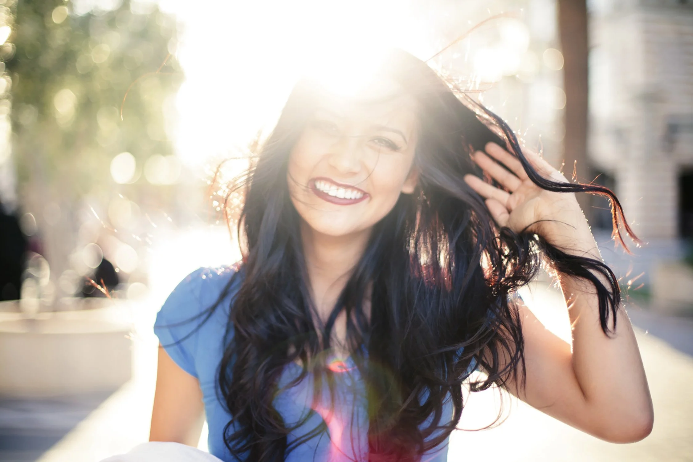 With a bright sun flare in camera and and behind the high school senior, Ashley. This is an up close image of Ashley trying to control her hair in the wind and smiling big with teeth.