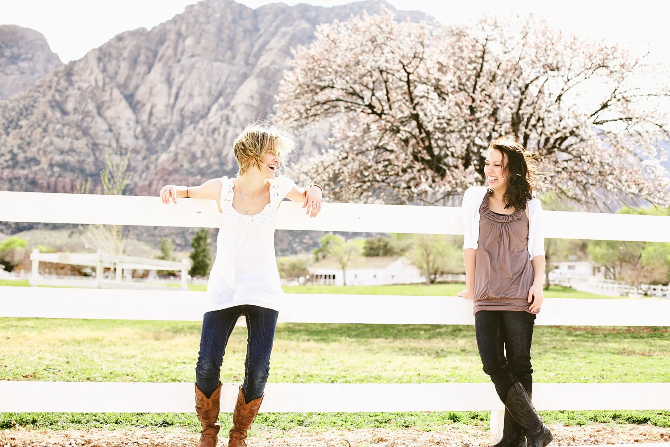 Two friends, young women, laughing looking at each other,  leaning up against a tall white wood fence with a big pink blooming tree and the mountain close behind it, at Spring Mountain Ranch