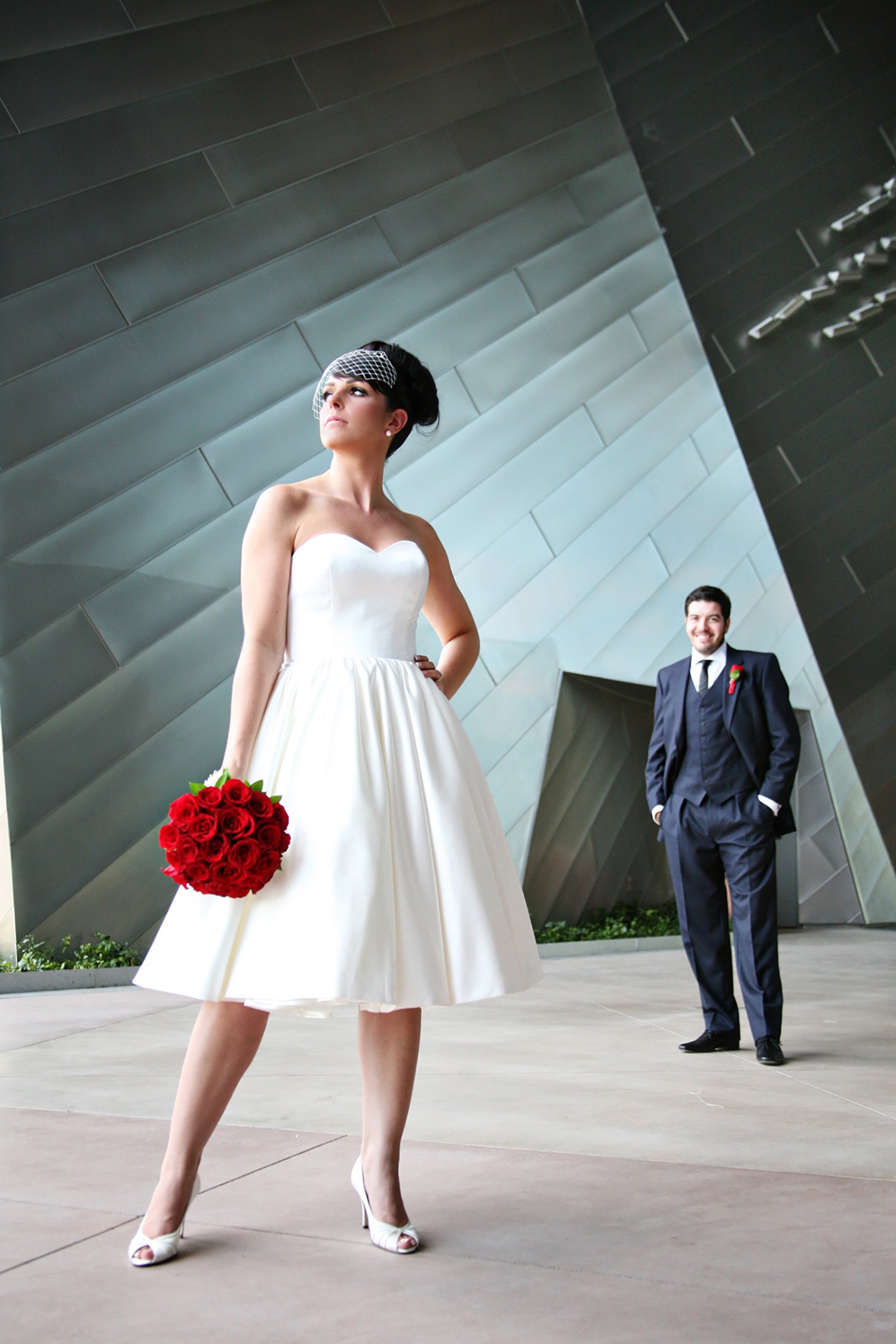 A newly married couple are standing in front of a matalic angled wall. The bride is in the foreground looking away from the camera, in a knee length sleeveless wedding dress holding a red bouquet of roses. The groom is in the background smiling