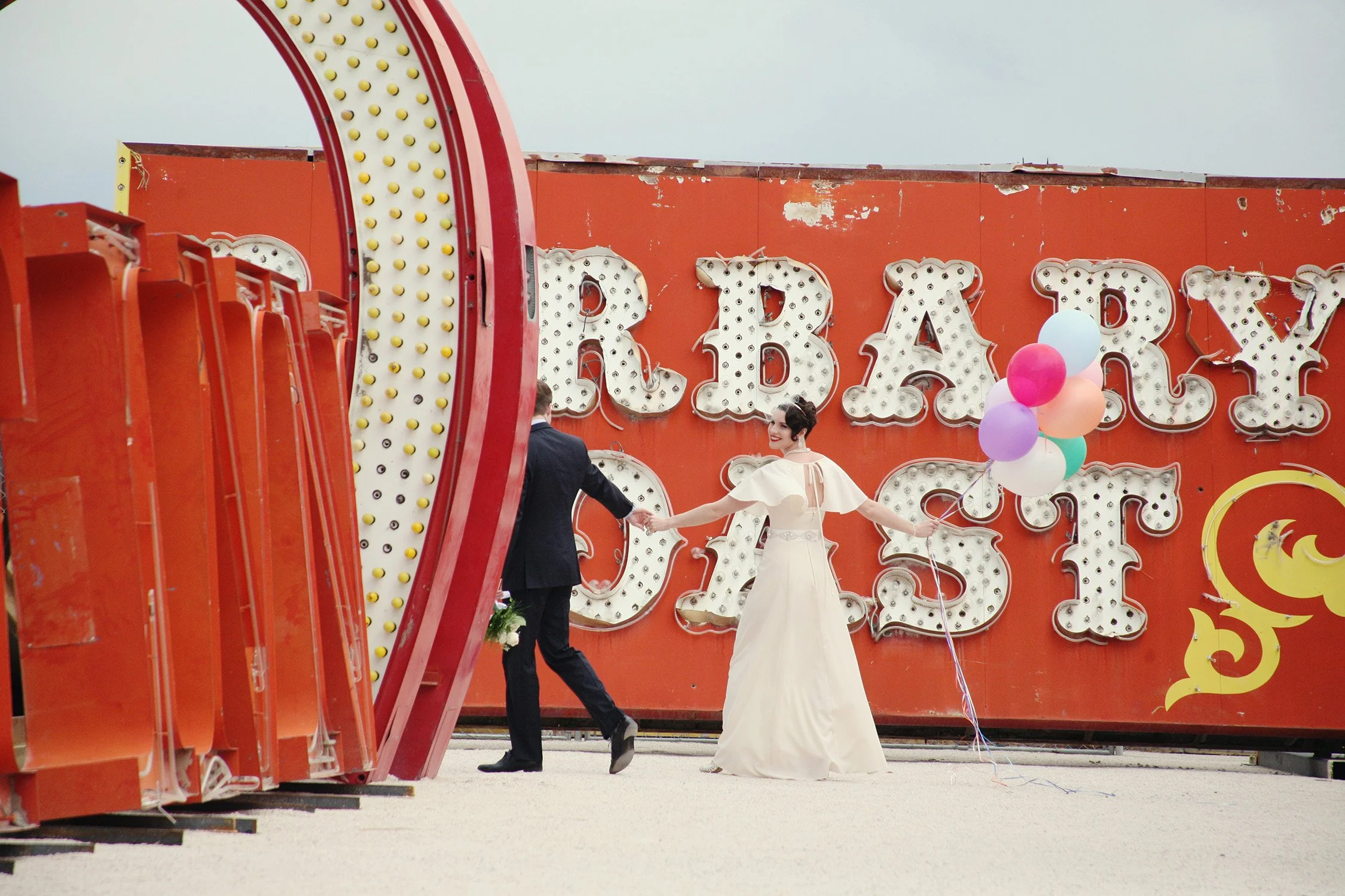Newlywed couple walking out of picture surrounded by decommissioned Vegas neon signs, bride is holding a bunch of balloons