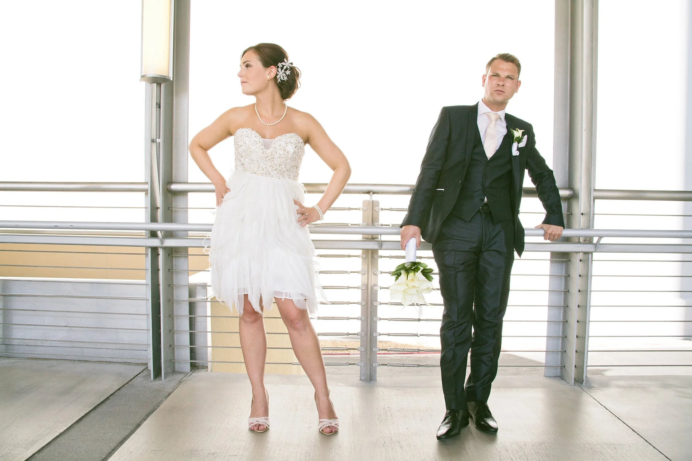 Newlywed couple in a suit and wedding dress, bride is looking away from camera hands on hips, groom is holding flowers leaning against railing looking at camera