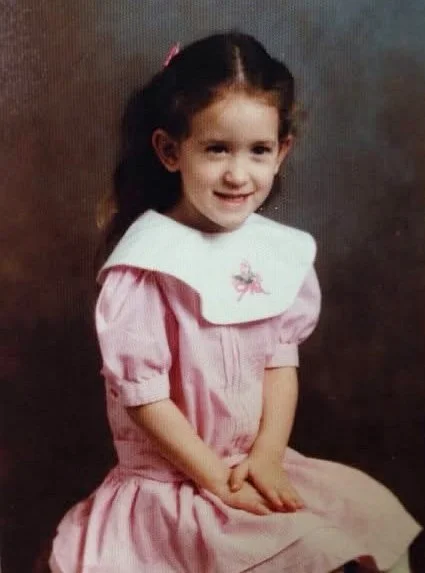 A four or five year old Lori in a pink and white dress, sitting with her hands in her lap and looking just off camera in a studio setting.