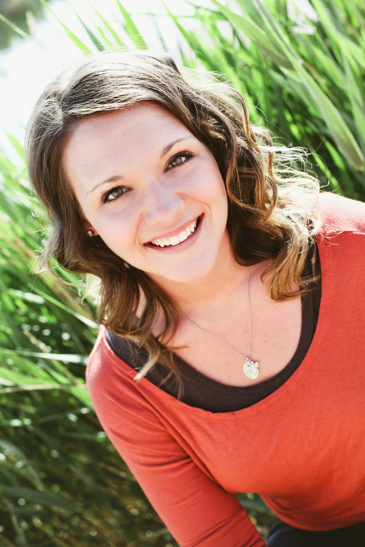 an up close image of a high school senior, she is smiling at the camera with tall green grass behind her. She is wearing an orange and brown shirt.