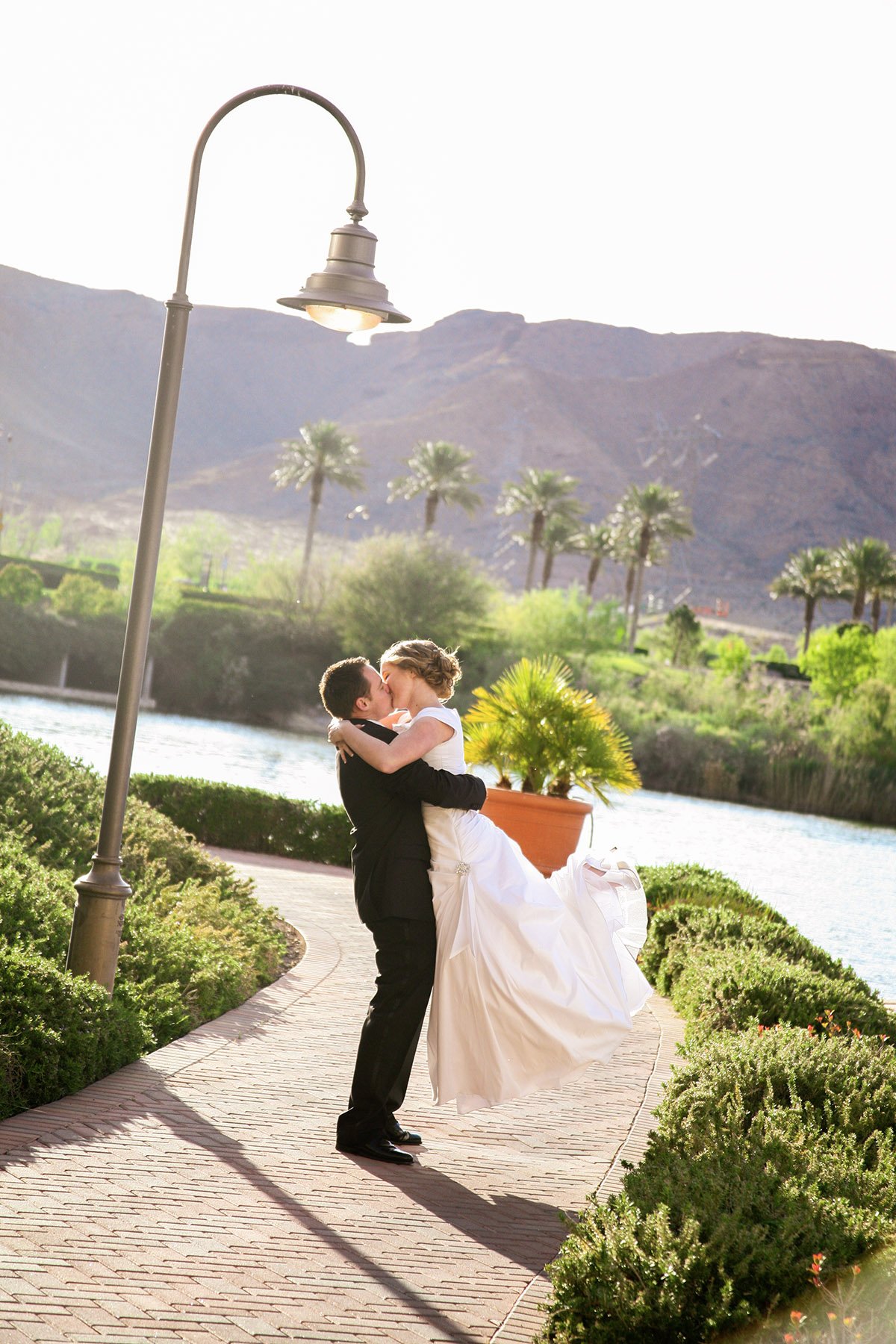 a newly wed couple kissing with the groom raising the bride off the ground, on a path lined with plants and a river in the background at Lake Las Vegas