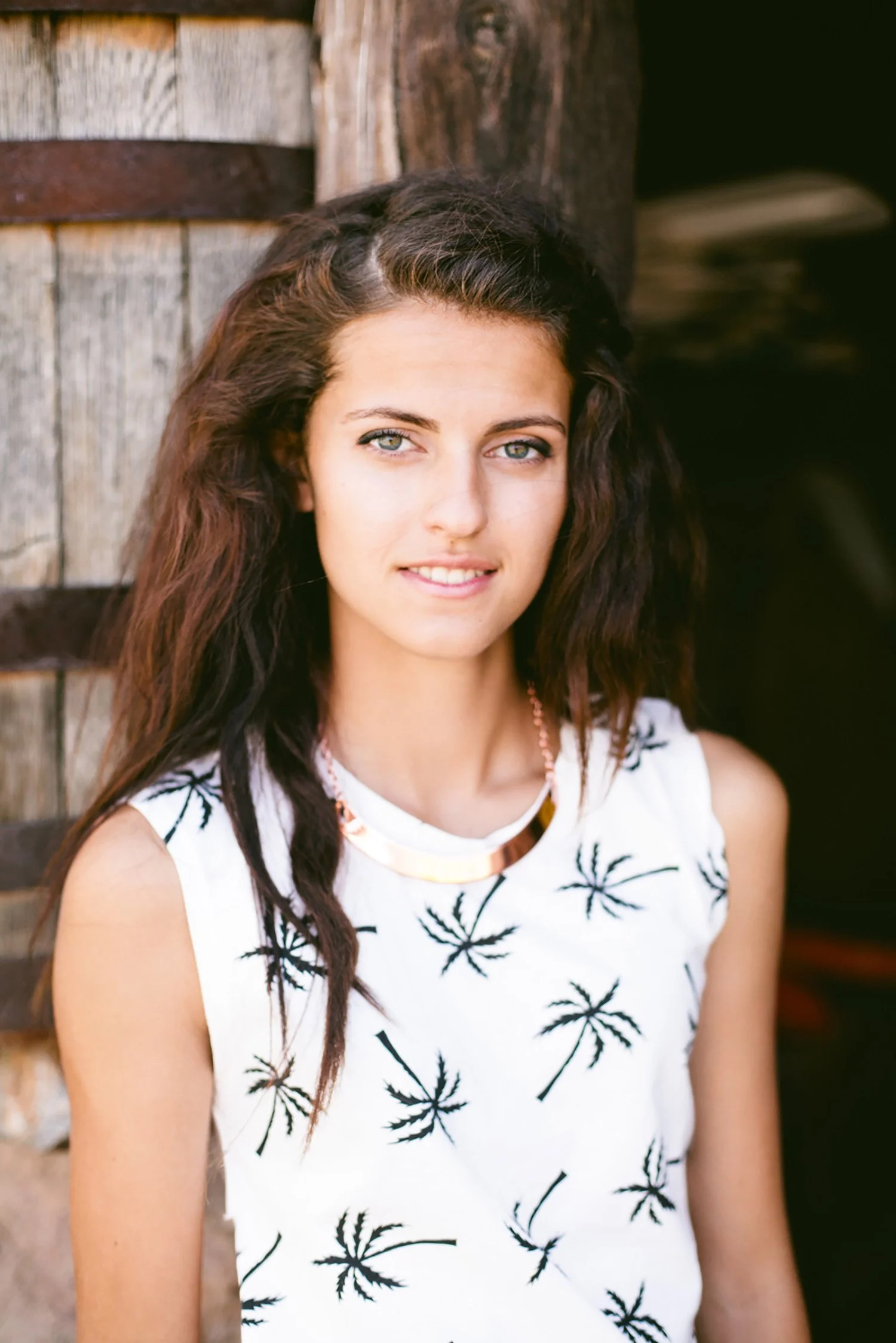 smiling and leaning up against a wood wall, a high school senior has long brown hair, and bright hazel eyes. 