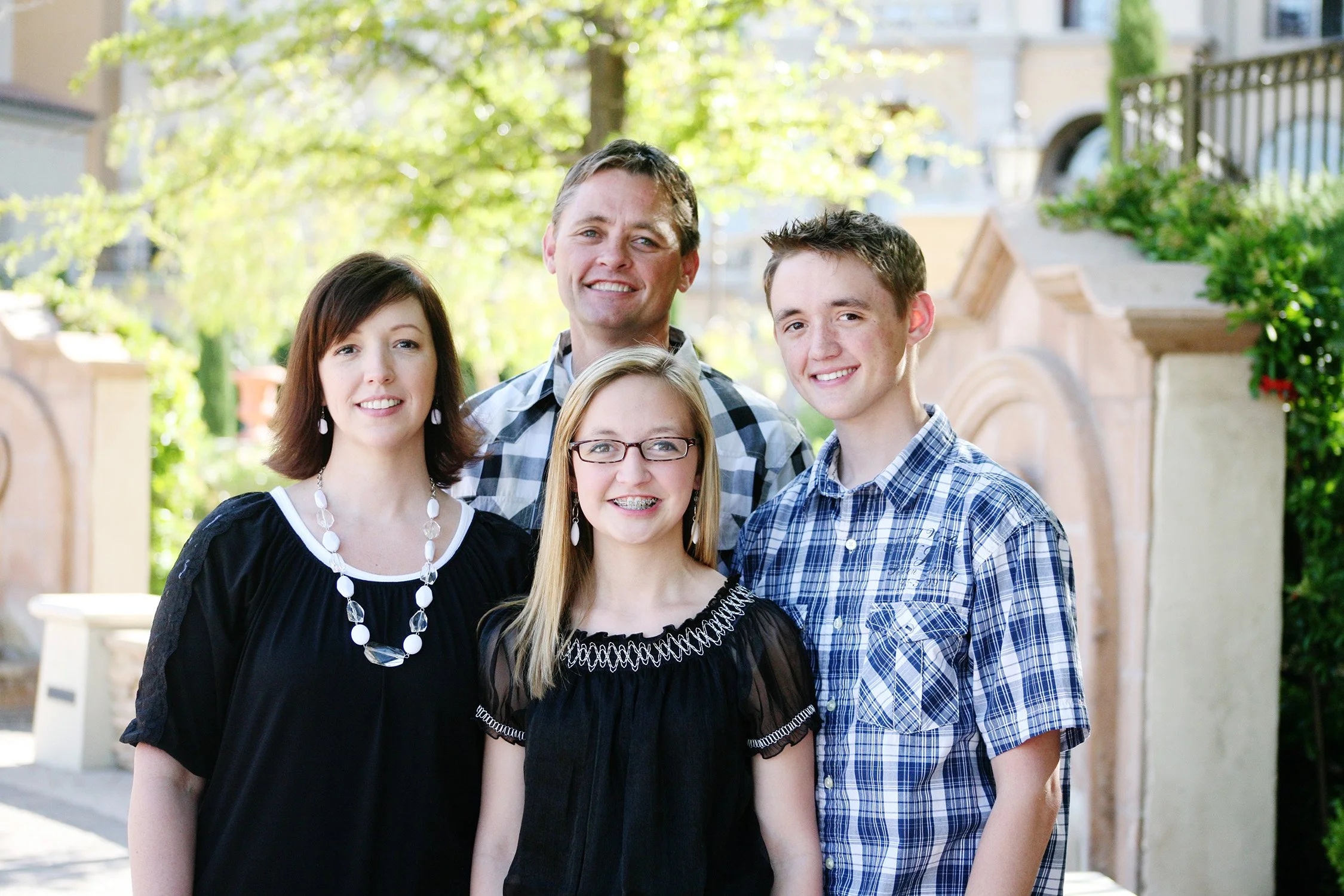 A family of four grouped close together, smiling at the camera with mom, dad and a older son and younger sister, a tree and brick feature wall are in the background.
