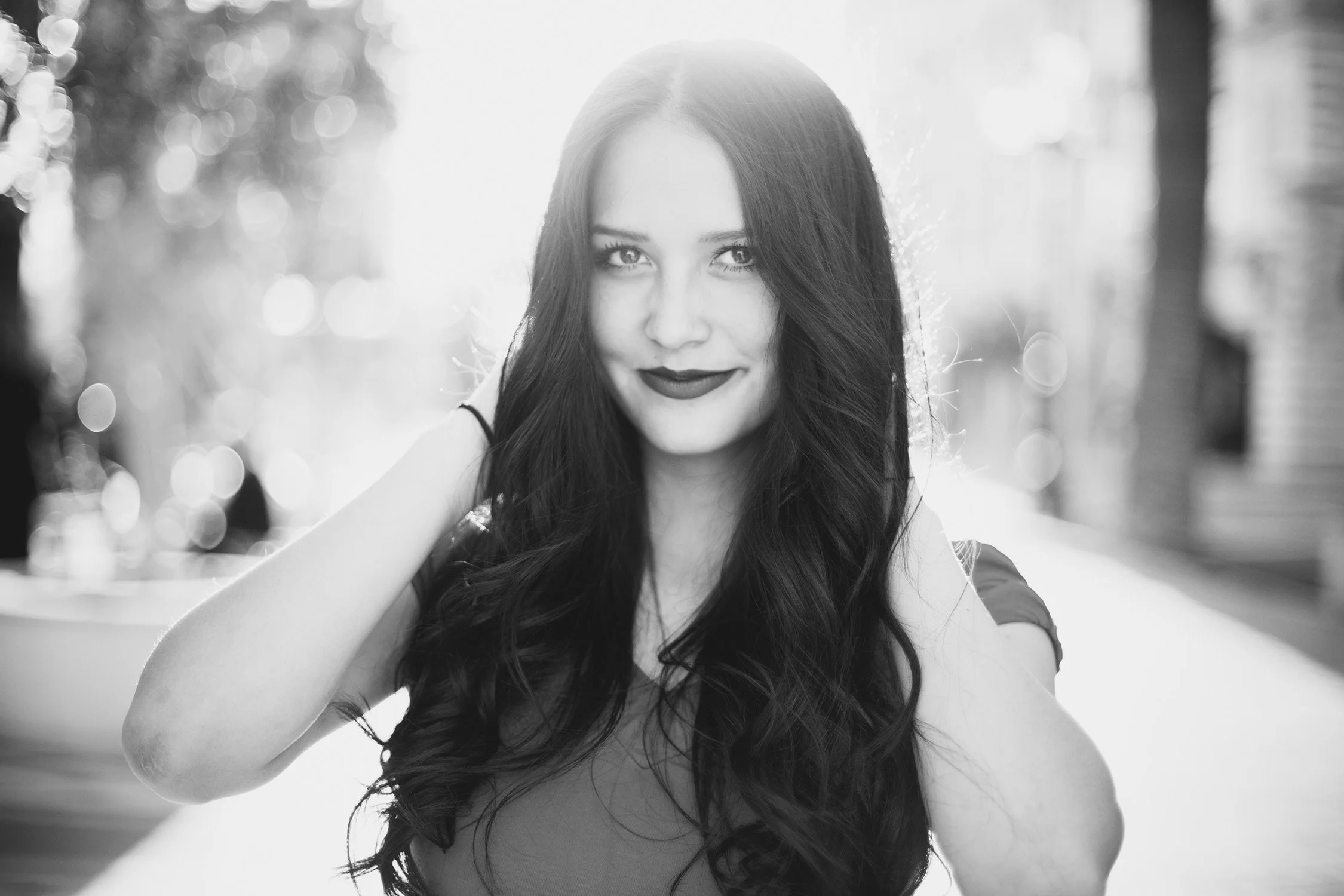 a black and white photo of high school senior, Ashley. She has her hands up near her hair and she is smiling at the camera with her mouth closed.