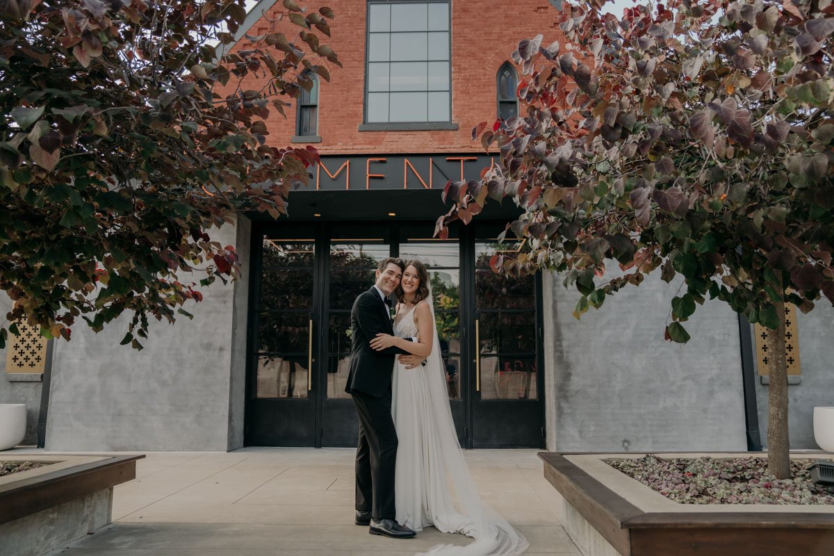 Couple embracing in front of a building with trees and sign reading "MENT," dressed in formal attire.