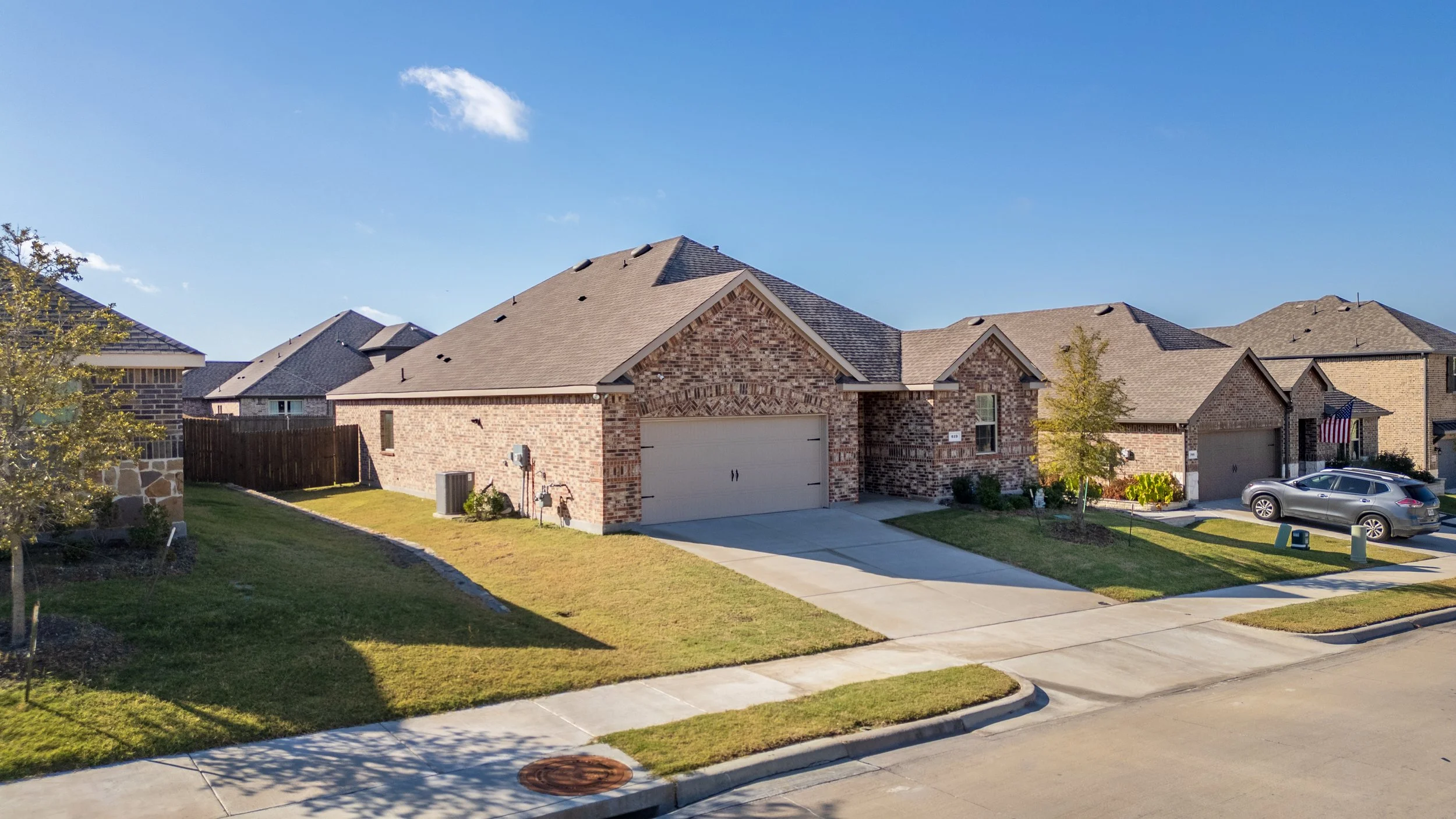A suburban neighborhood with brick houses, one with a two-car garage and a gray SUV parked in the driveway, green lawns, and a tree, under a clear blue sky.