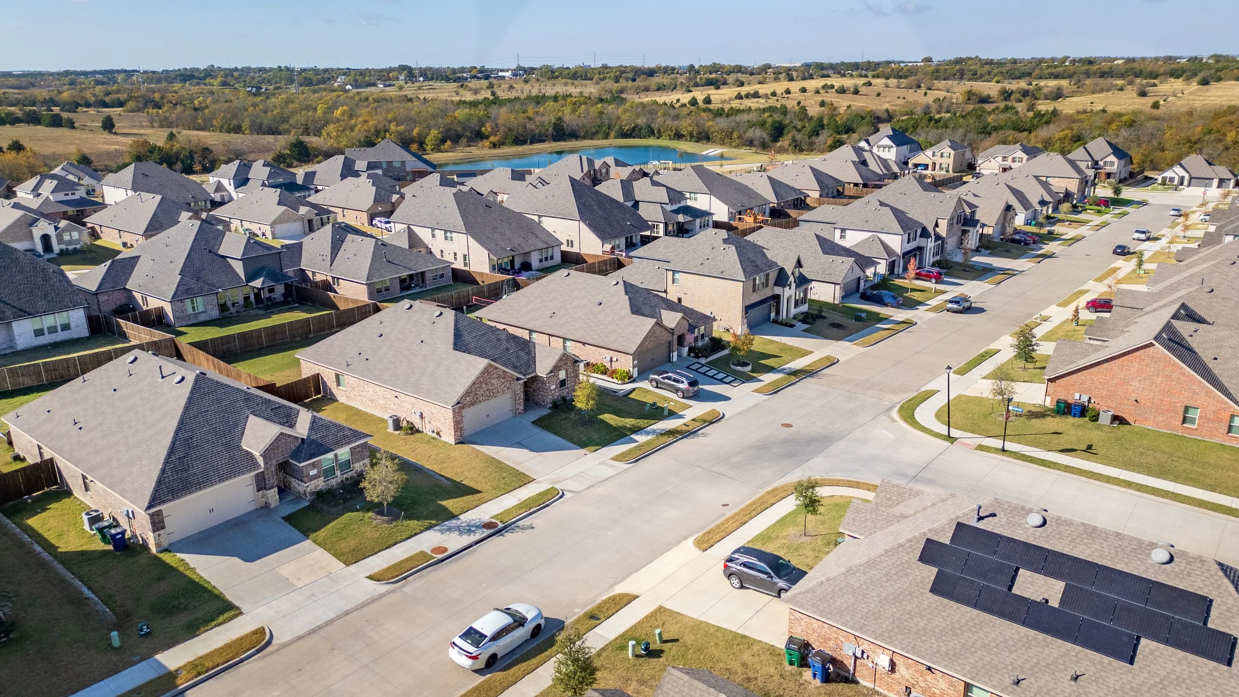Aerial view of a suburban neighborhood with rows of single-family houses, manicured lawns, cars parked along the streets, and a pond in the background under a partly cloudy sky.