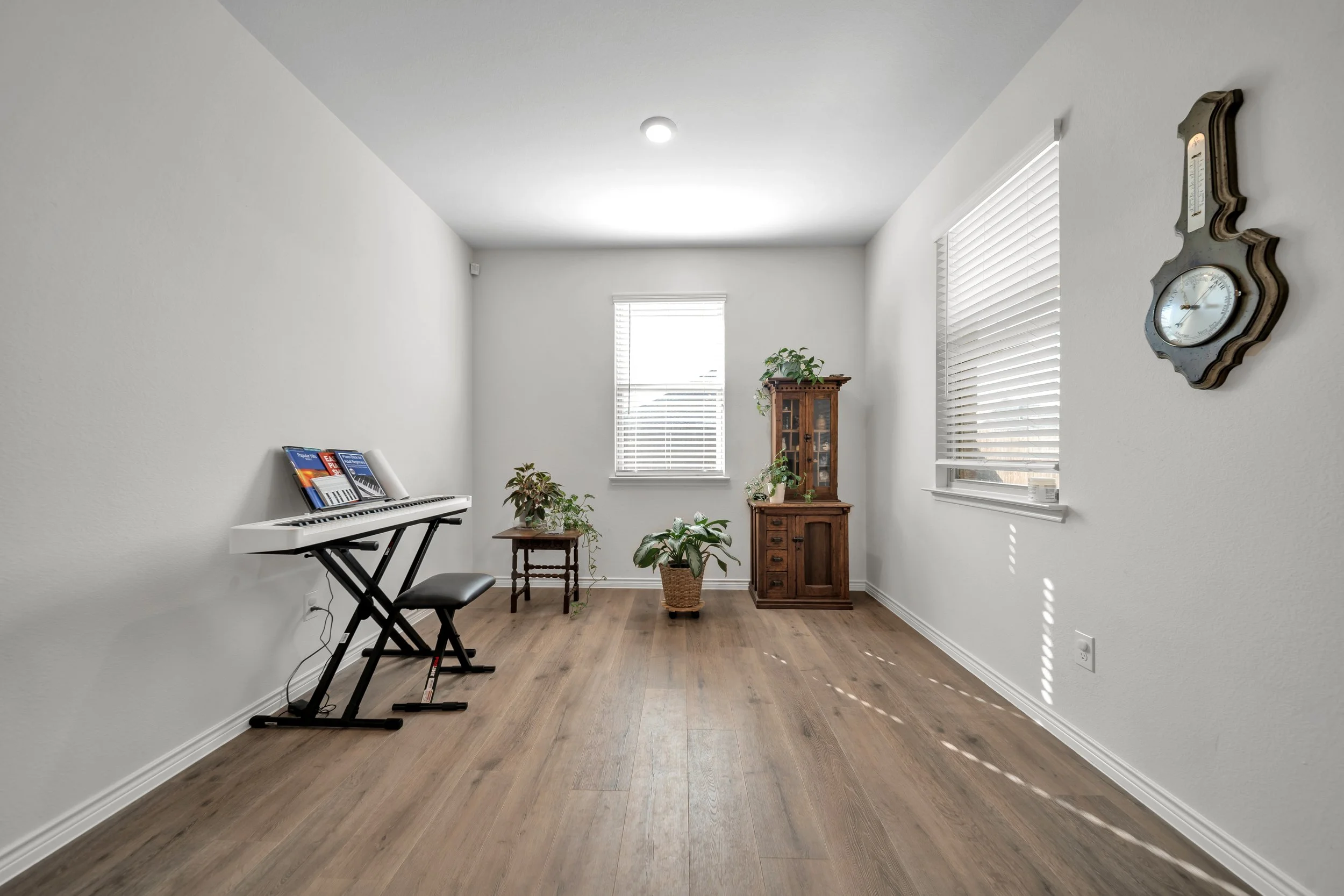 A minimally furnished room with white walls, a wooden floor, and two windows with white blinds. The room contains a digital piano with sheet music, a small wooden table with a plant, a larger wooden cabinet with plants on top, and a wall clock.