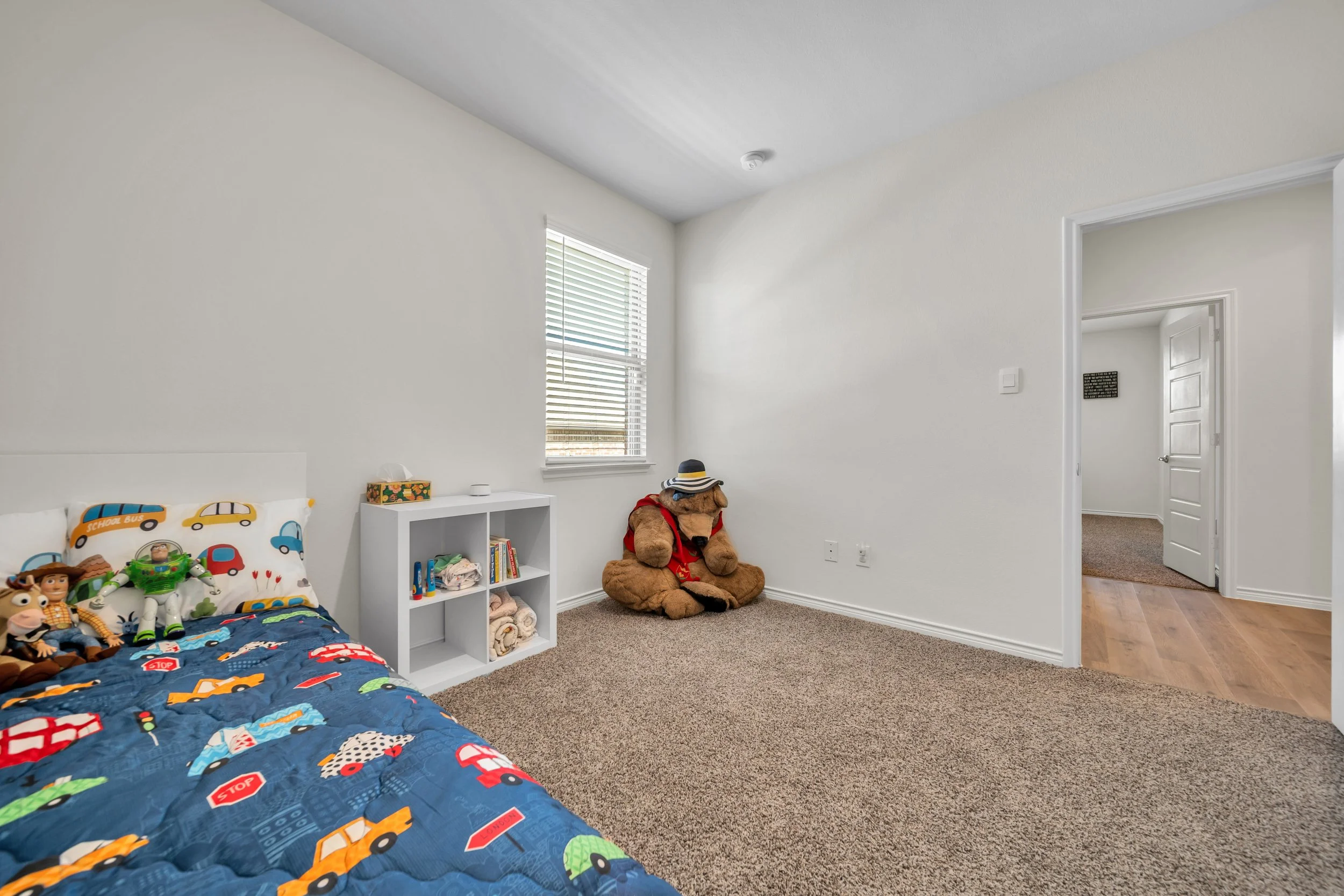 Children's bedroom with a bed decorated with vehicle-themed bedding and plush toys, a white cubby shelf with books and folded blankets, a large teddy bear wearing a hat and red shirt sitting against the wall, beige carpet, white walls, and a window w