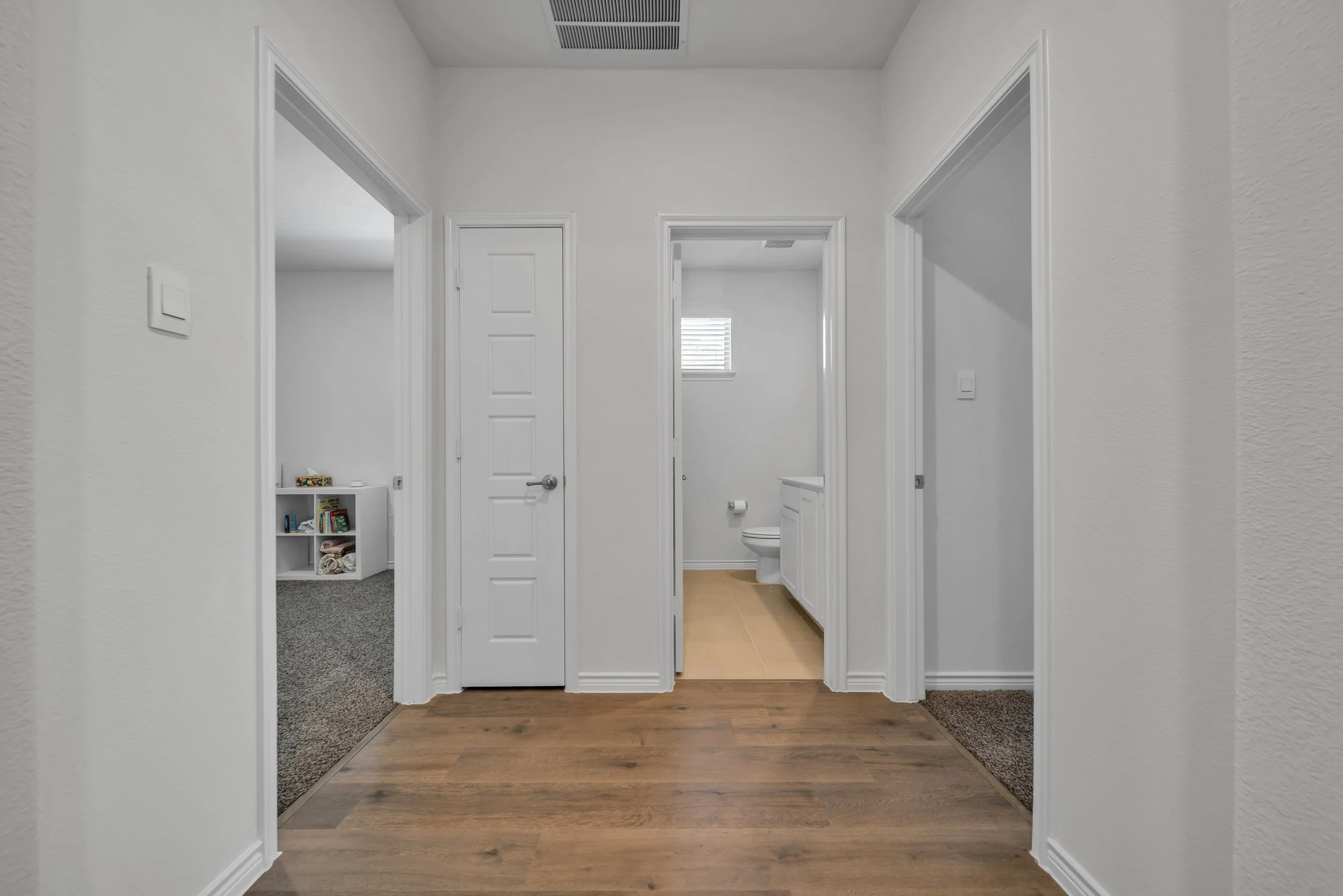 Interior hallway view showing three doorways, one leading to a room with carpet, one to a bathroom with a toilet and small window, and one to a space with a closet. The floor has wood and carpeted areas, white walls, and a ceiling vent.
