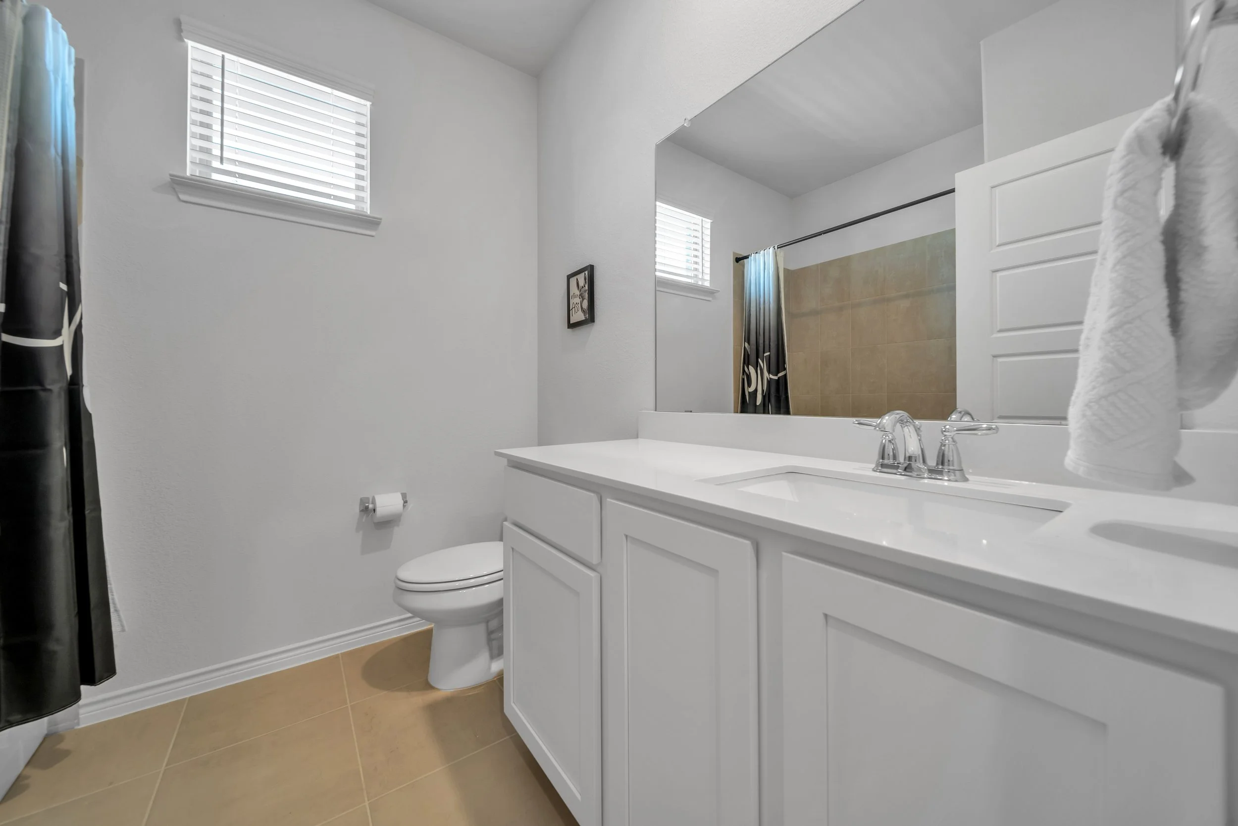 A clean bathroom with white cabinets, a large mirror, a white toilet, beige tiled flooring, a small window with blinds, and a shower with a curtain.