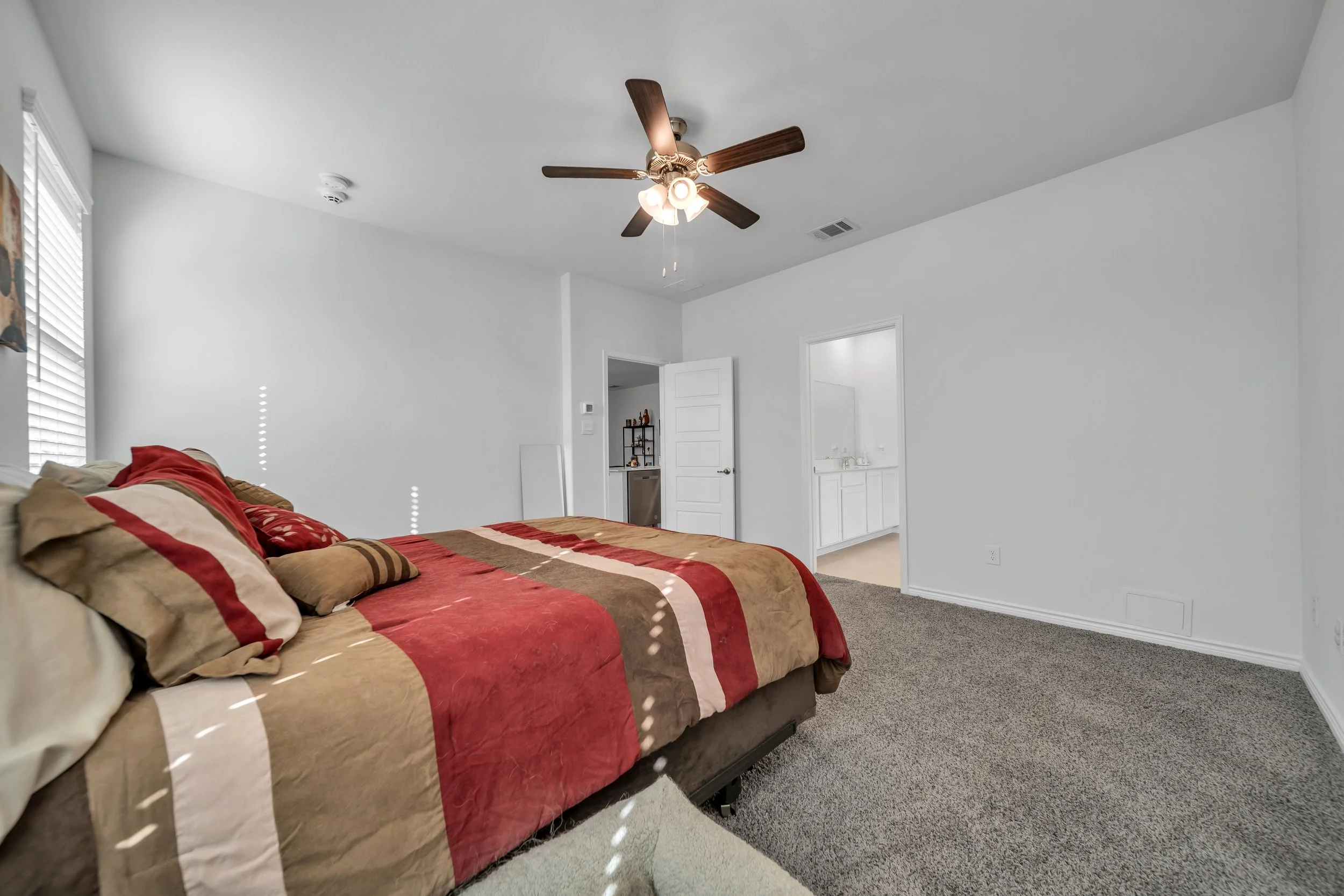 Bedroom with a bed covered in a brown, red, and white striped comforter and pillows, white walls, a ceiling fan, carpeted floor, and a doorway leading to a bathroom.