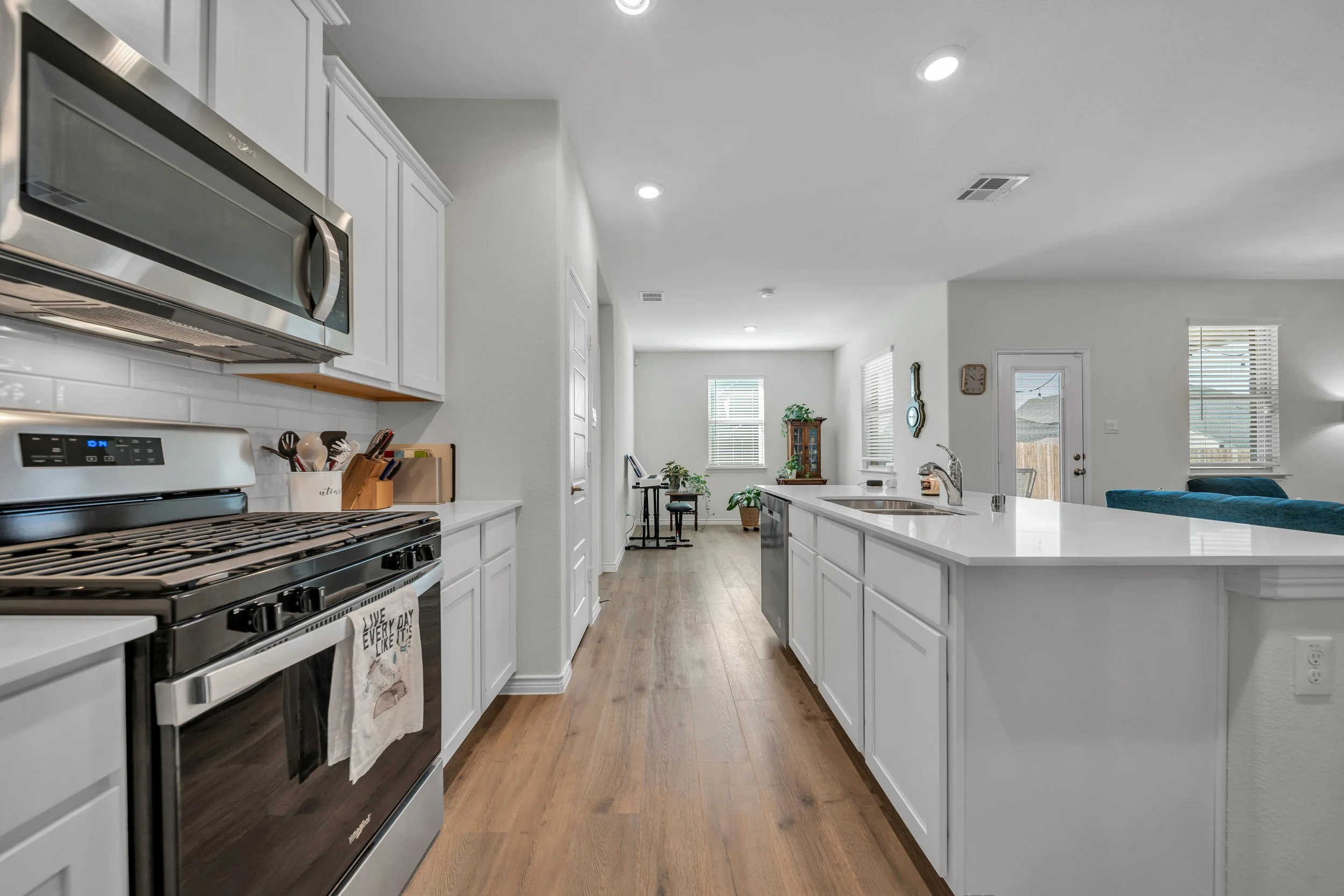 Open kitchen with white cabinetry, stainless steel appliances, a white countertop island, hardwood floors, and a view into the living and dining area with windows and furniture.