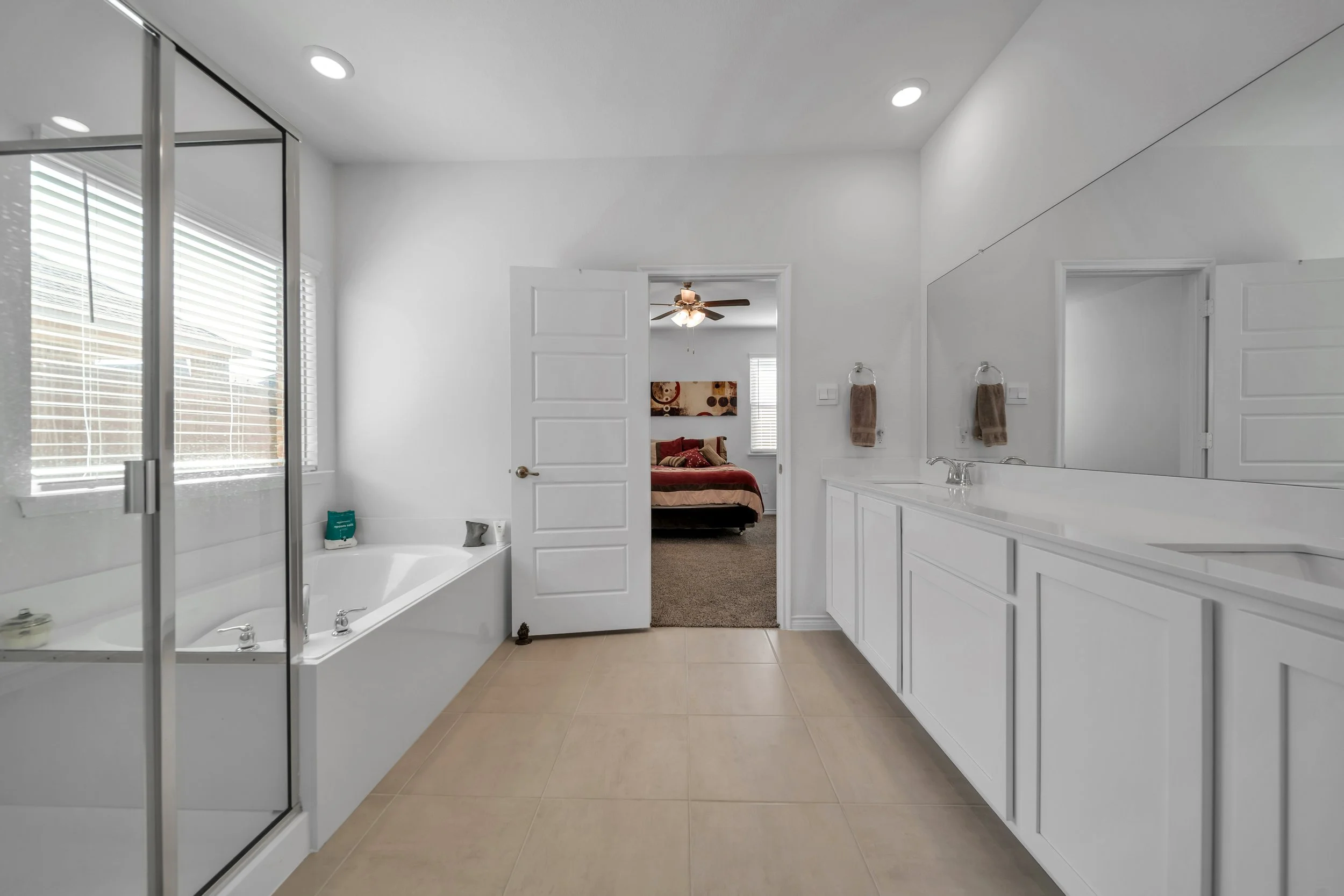 Modern bathroom with beige tiled floor, white cabinets and walls, large mirror, glass shower enclosure, bathtub, and view into a bedroom with a bed, ceiling fan, and artwork.