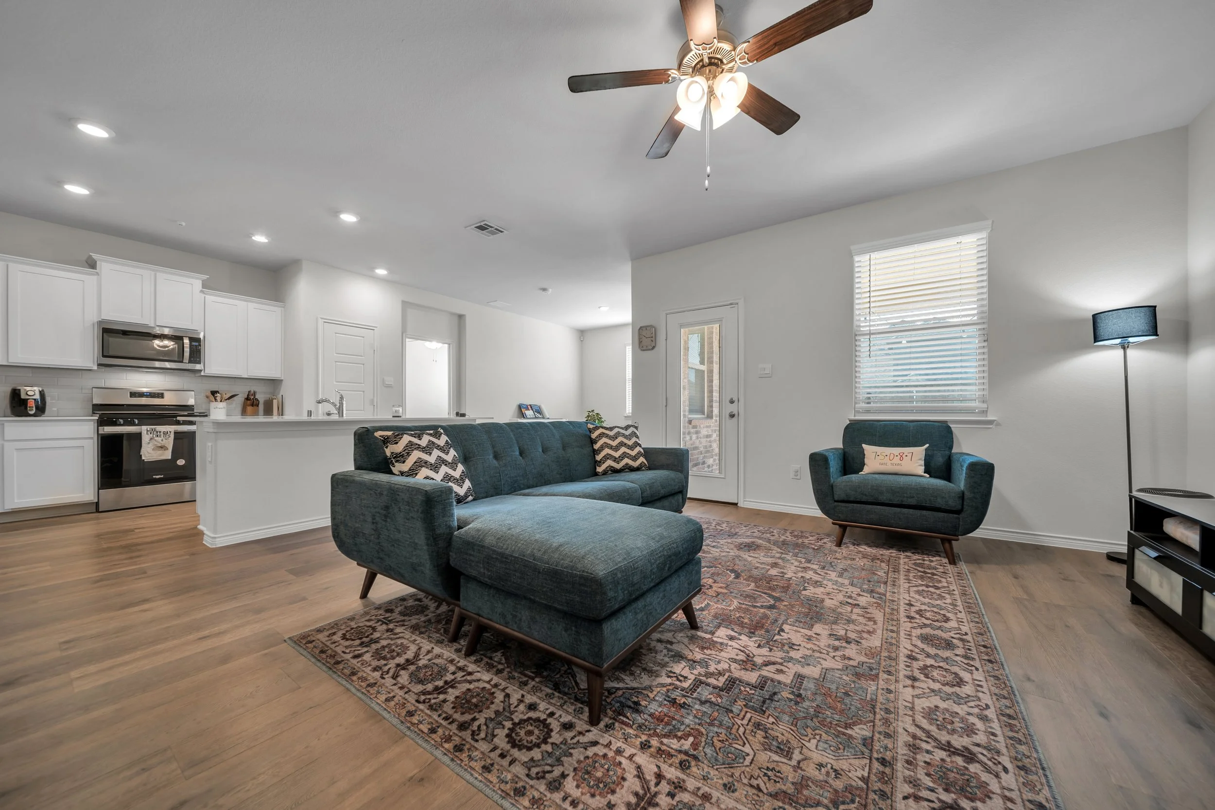 Living room with blue seating, decorative rug, and kitchen in the background, ceiling fan, window, and door.