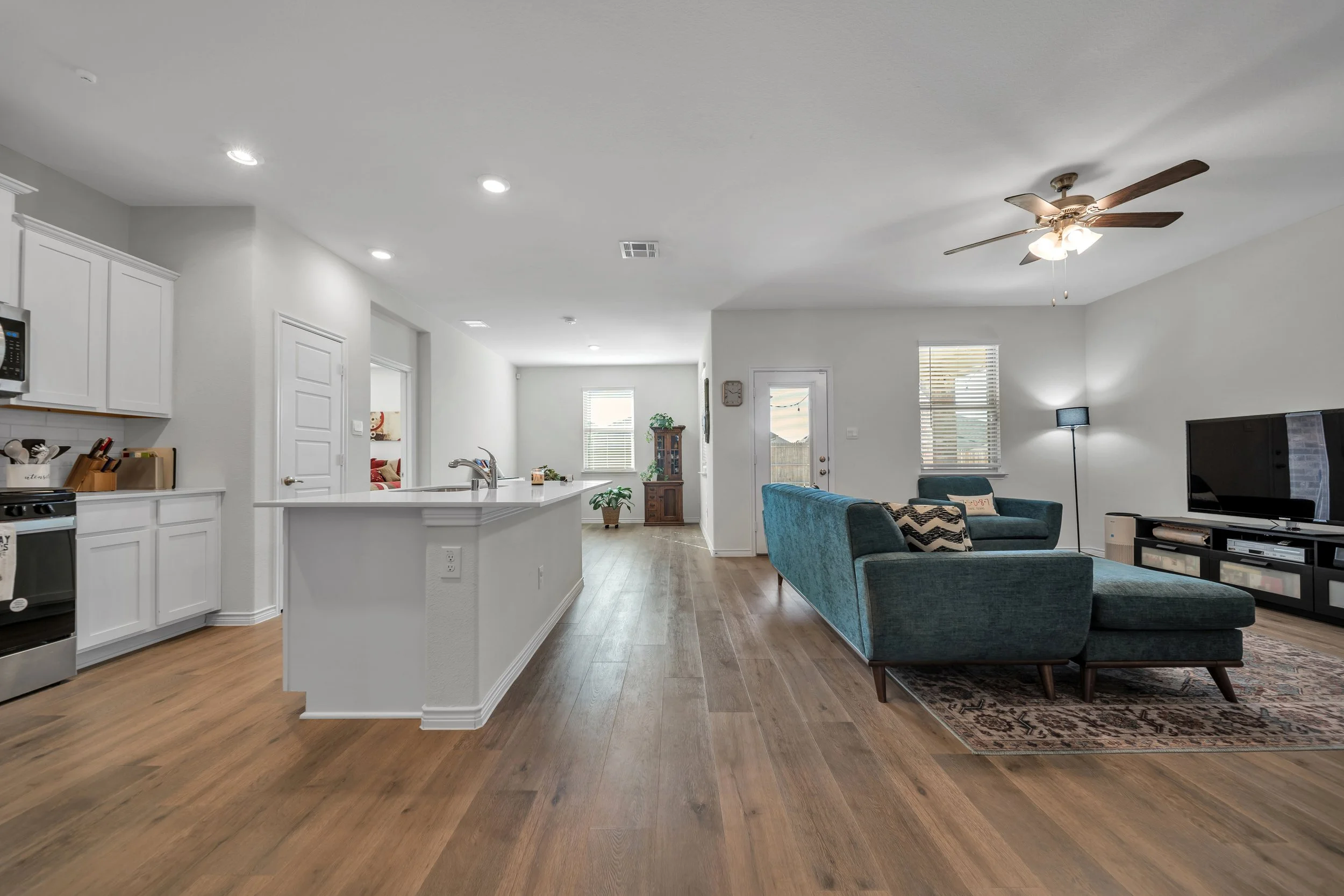 Open-concept living room and kitchen with hardwood floors, white walls, a ceiling fan, a teal sectional sofa, a flat-screen TV, and large windows providing natural light.