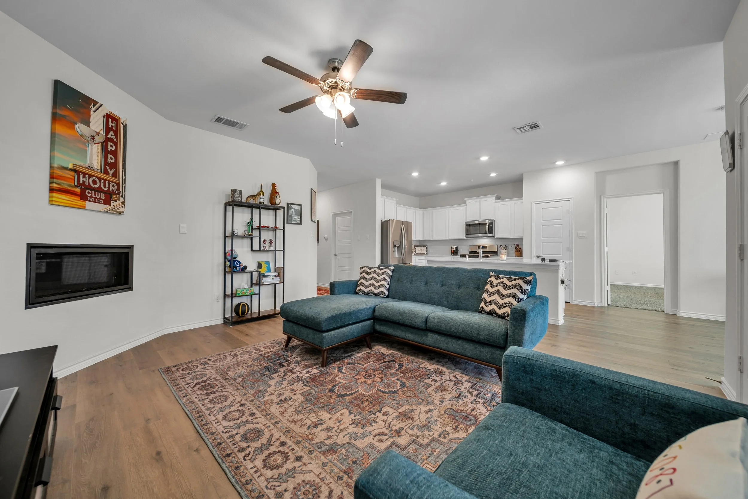 Living room with a teal sectional sofa, patterned throw pillows, a patterned area rug, a black open shelving unit, a wall-mounted electric fireplace, white walls with a colorful "Happy Hour" wall art, a ceiling fan, and an open kitchen with white cab