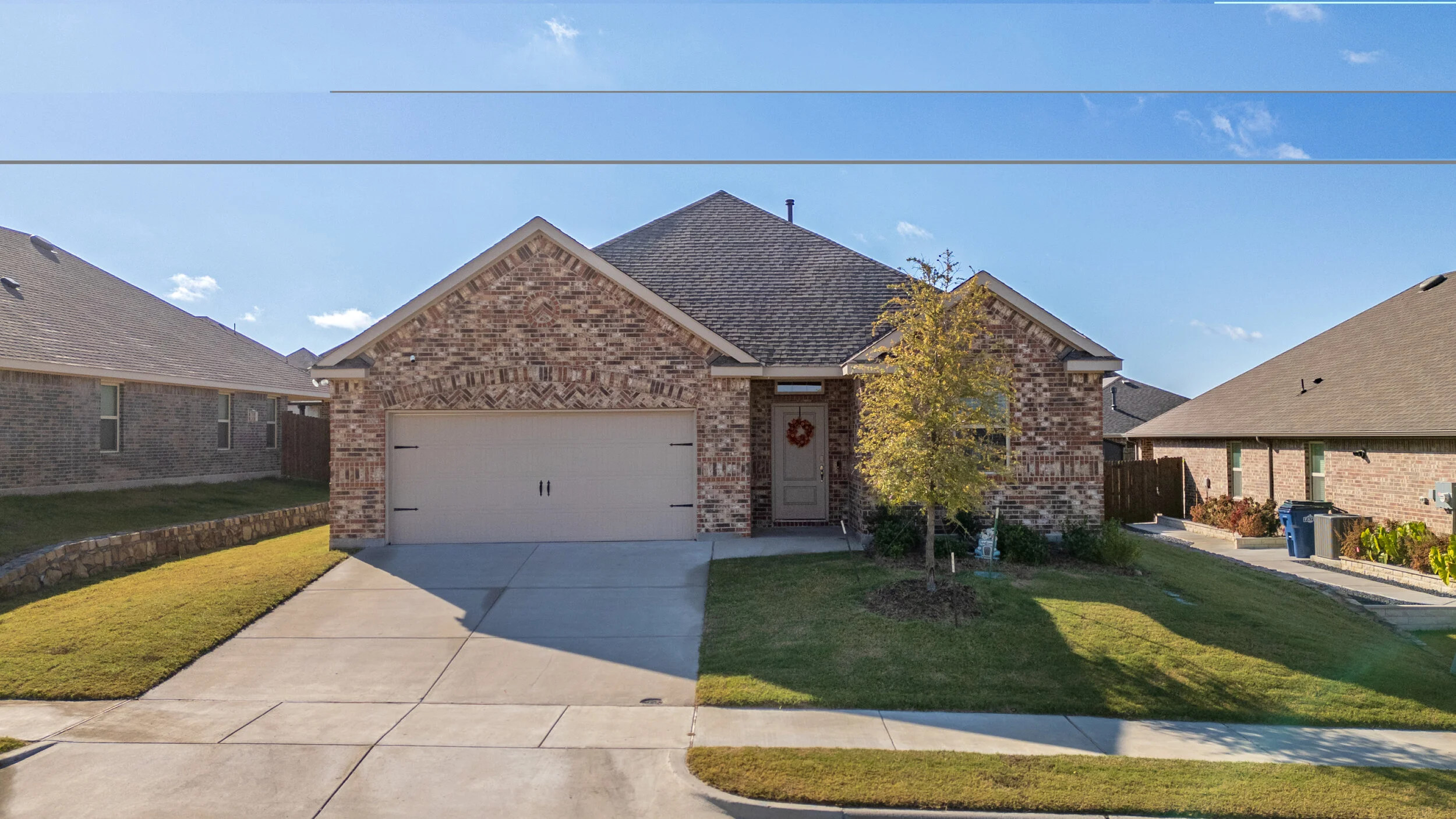 Front view of a brick house with a two-car garage, a small front lawn, a tree, and neighboring houses on a sunny day.